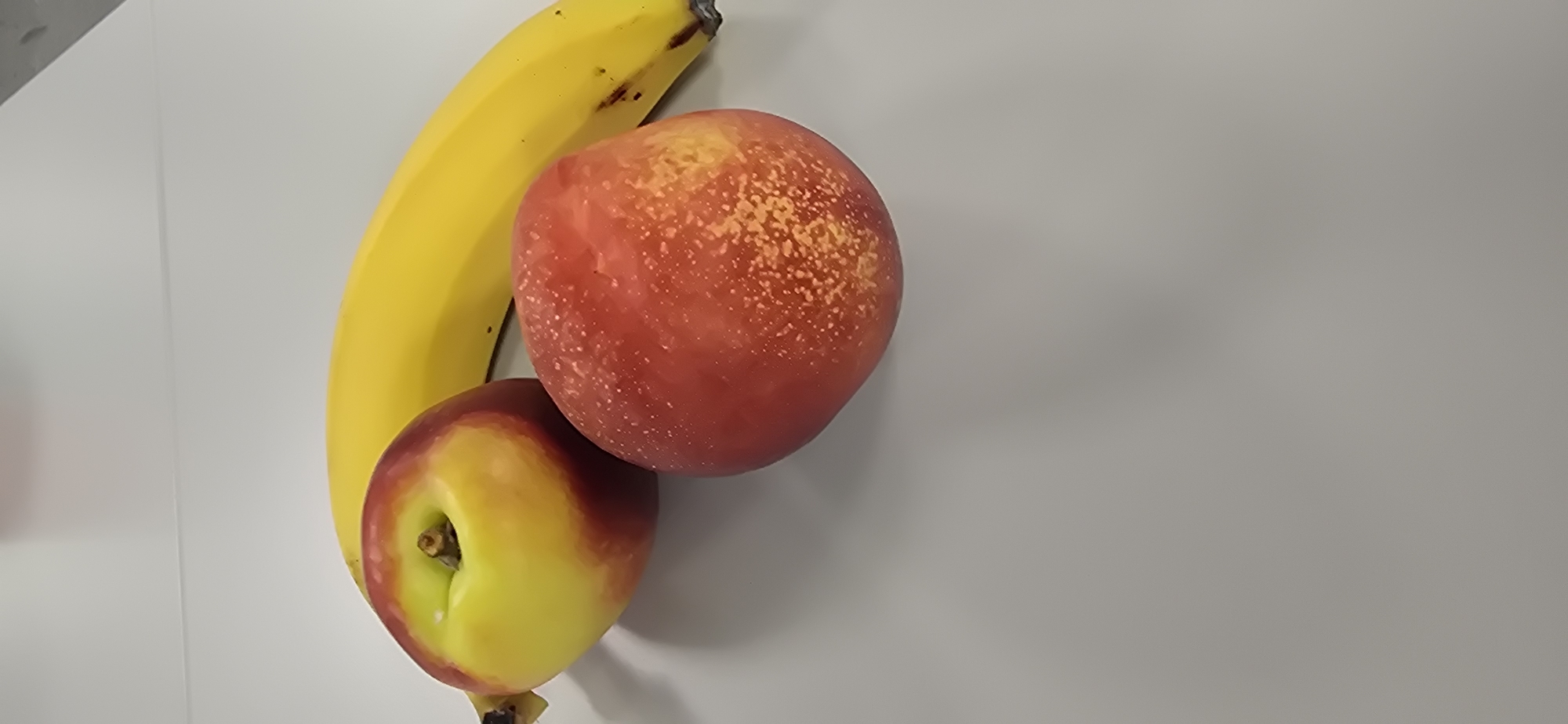 A top-down view of a yellow banana, a red and yellow nectarine, and a red peach are arranged on a plain, light gray surface. The banana is positioned vertically on the left side of the frame, with its stem pointing towards the top. The nectarine is placed below and slightly to the right of the banana, while the peach is situated above and to the right of the nectarine, partially overlapping the banana. The fruits are the sole subjects in the image. There are no people, text, or specific location cues visible. The lighting suggests an indoor setting, possibly a countertop or table, with a soft, diffused light source. No weather or time of day can be determined. The image is a still life of the fruit.