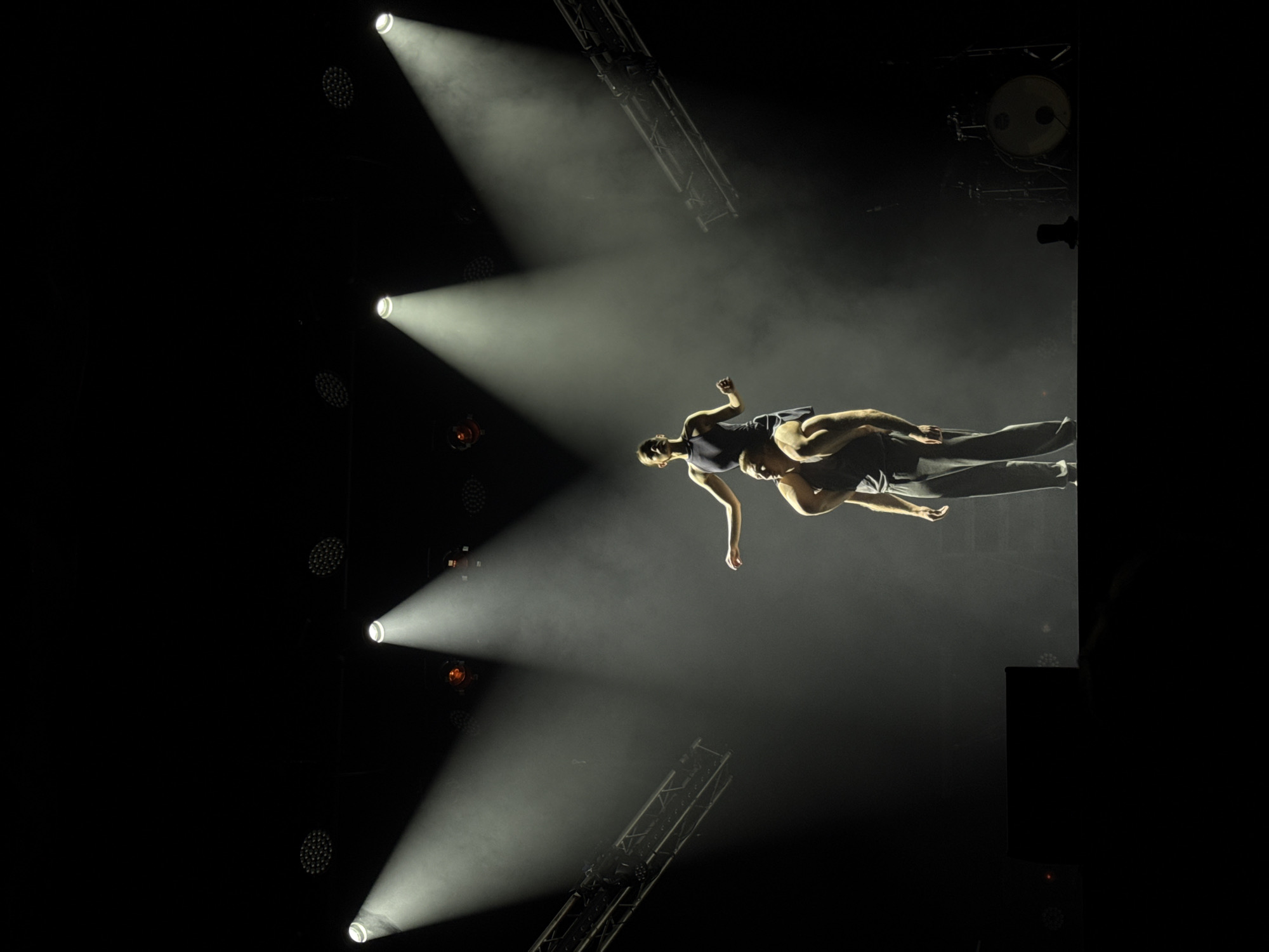 The image captures a dramatic performance on a dimly lit stage, illuminated by three bright spotlights casting shafts of light downwards. In the foreground, a male dancer is positioned, supporting a female dancer who is seated on his shoulders. The male dancer is facing forward, his muscular build evident in his bare arms. He wears dark, loose-fitting pants. The female dancer is facing to the left, her arms extended outwards, her posture suggesting poise and balance. She is wearing a dark, sleeveless leotard or dress.
The background is largely dark, with stage rigging visible on the left and right sides of the frame. The lighting creates a sense of depth and theatricality. On the far right of the stage, a drum kit is visible, suggesting the presence of live music accompanying the performance. The overall atmosphere is one of artistic expression and physical prowess. The location is not specified in the image.