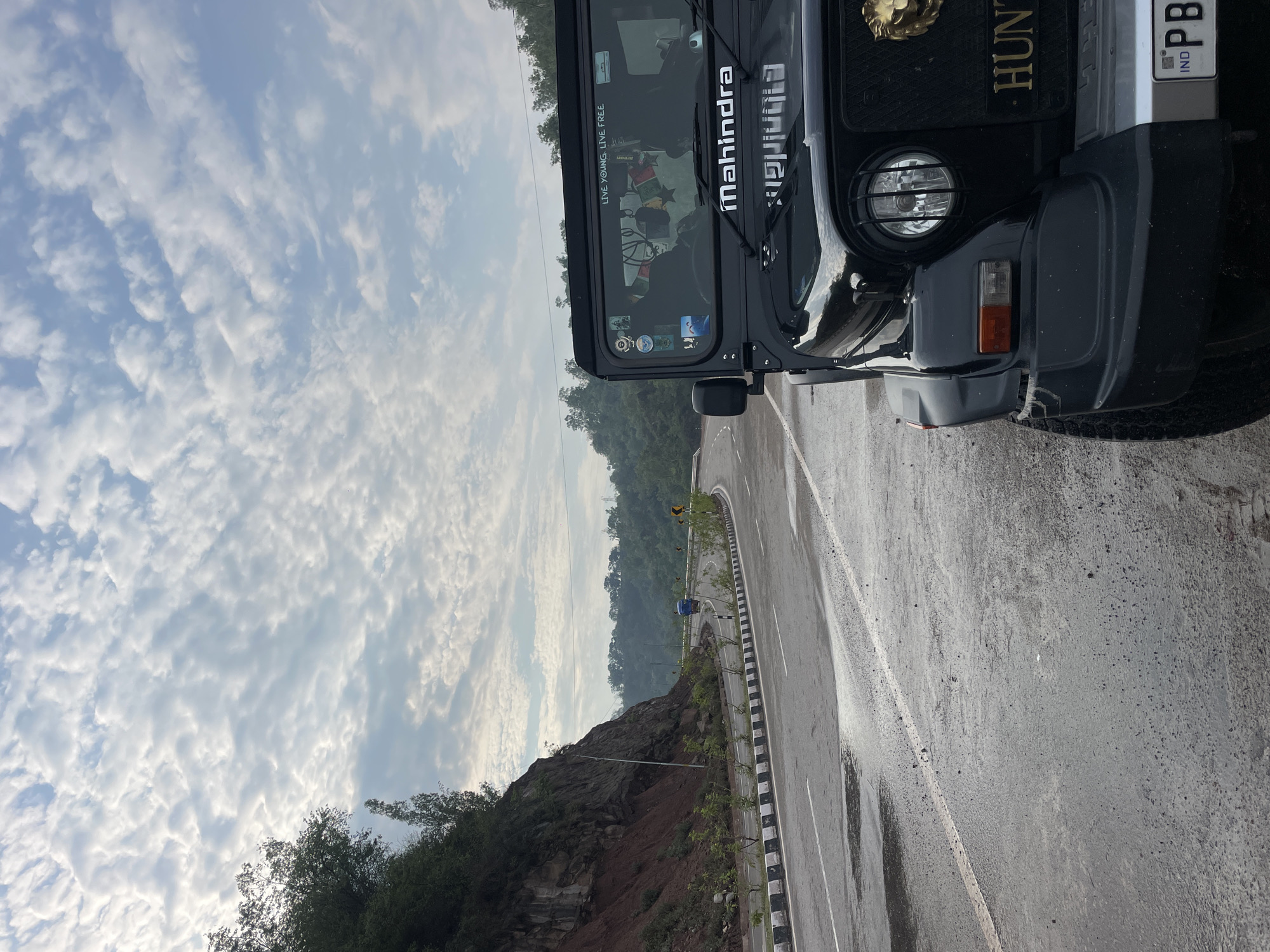 An outdoor, eye-level shot captures a black Mahindra Thar SUV parked on the side of a wet, winding road in Trilokpur, India. The vehicle's front end, including its headlights and a golden lion emblem on the grille, is prominent in the foreground. The road is lined with a black and white striped barrier and features white lane markings. To the left, a steep, rocky embankment covered in vegetation rises above the road. In the distance, the road curves through lush green hills under a sky filled with scattered, fluffy white clouds. The weather appears overcast, and the road's wetness suggests recent rain. No people are visible in the image.