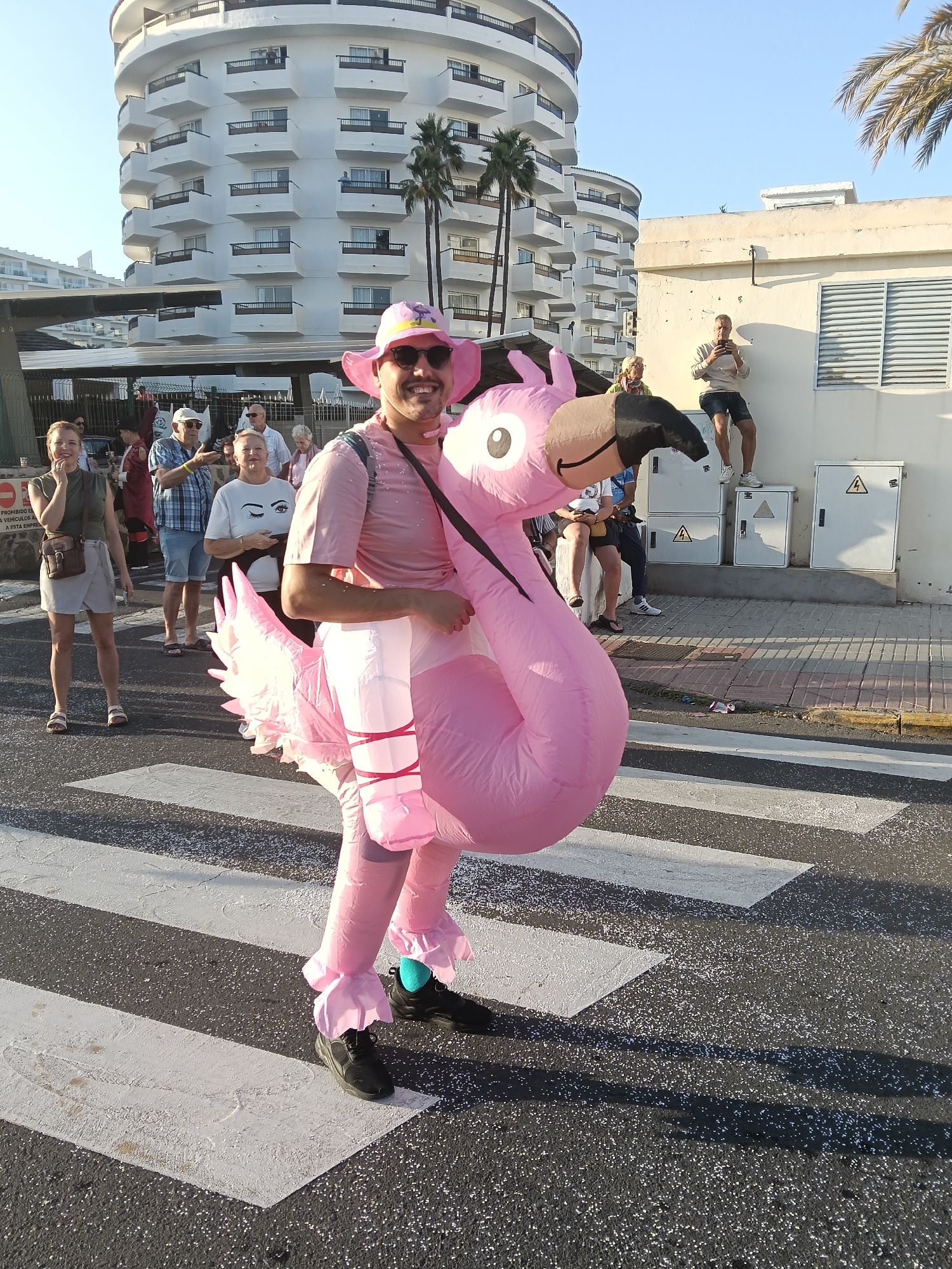 A man dressed in a pink flamingo costume with a pink hat and sunglasses stands on a zebra crossing in San Bartolomé de Tirajana, Spain. He is surrounded by other people, including a woman with a brown bag, a man in a blue plaid shirt, and a woman in a white shirt with a graphic of eyes. Behind them is a tall white building with curved balconies and palm trees. To the right, a man stands on top of a set of electrical boxes, taking photos. The road is littered with small, reflective pieces of trash.