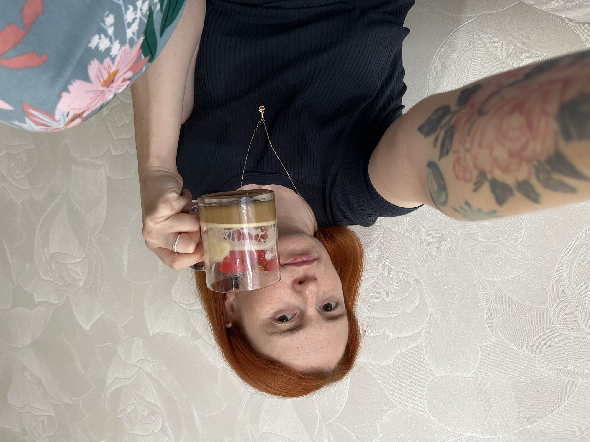 An adult female, approximately in her late 20s to early 40s, is captured in a head-and-torso selfie, likely taken indoors in the city of Chaikovsky, Russia. The subject has shoulder-length, coppery-red hair and a light complexion marked with scattered freckles across her face. Her expression is neutral, and she looks directly into the camera. She wears a dark-colored, possibly navy or black, short-sleeved ribbed top and light blue or teal pants with a large floral pattern featuring pink, white, and green elements. Around her neck is a thin, gold-colored chain necklace with a small, teardrop-shaped pendant containing a clear stone. On the ring finger of her left hand, she wears a thin, textured silver-colored ring. Her left forearm displays a tattoo sleeve, featuring a prominent large pink flower (possibly a peony) with green leaves, alongside darker, abstract shapes. She is holding a clear glass mug with her right hand; the mug contains a layered beverage consisting of a light brown liquid (likely coffee or a latte), topped with a white foamy layer, several red fruit pieces (resembling strawberries or raspberries), and lighter fruit slices (possibly banana). The background is an off-white or light cream wall covered in textured wallpaper, embossed with a large, repeating floral pattern depicting stylized rose-like blossoms and foliage.
