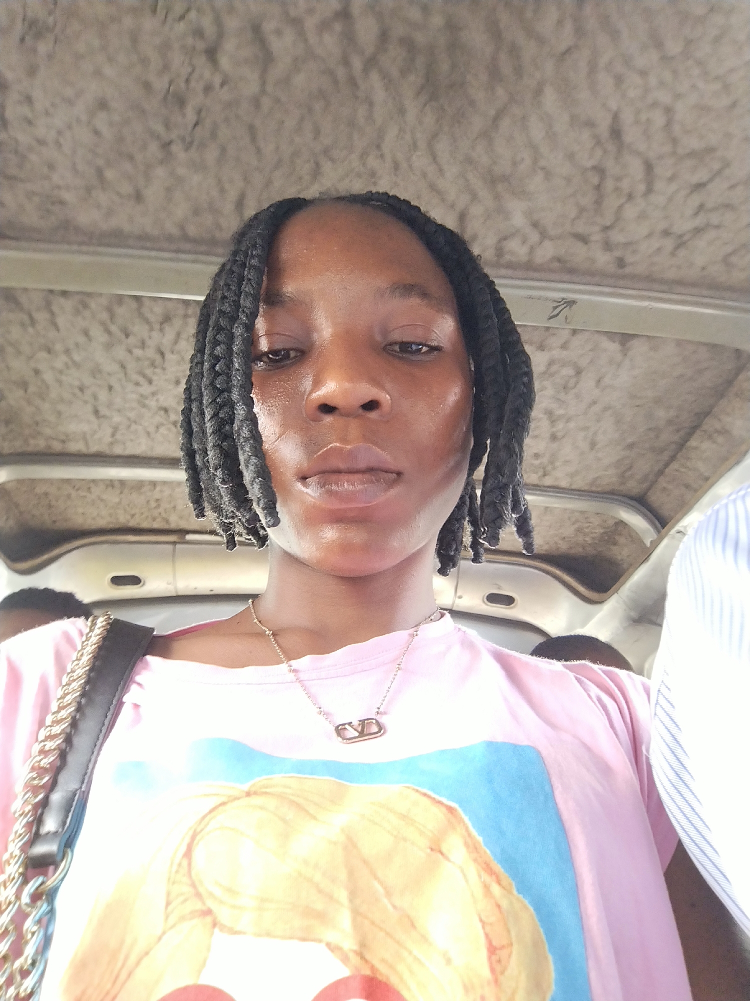 The image is a close-up, selfie-style shot of a young Black woman with braided hair, wearing a pink t-shirt with a graphic print and a gold necklace. She appears to be inside a vehicle, possibly a bus or van, with a textured, light-colored ceiling visible above her. The background includes parts of other passengers and the interior of the vehicle. The lighting suggests an indoor setting, possibly during the day. The image is taken from a slightly high angle, looking down at the subject. The quality of the image is moderate, with some graininess and a focus on the subject's face and upper body. There are no discernible landmarks or specific environmental cues that would pinpoint the location to Benin City, Nigeria, beyond the context provided. The overall impression is a casual portrait captured during transit.