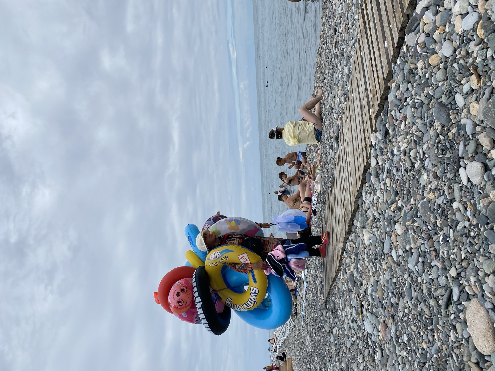 A vendor stands on a wooden walkway on a pebbled beach, carrying a large assortment of inflatable rings and other beach toys. The vendor is wearing a light-colored hat, a patterned shirt, dark pants, and red shoes. They are holding a white bag with blue trim in their left hand. Behind the vendor, several people are relaxing on the beach and in the water. The sea is calm with a cloudy sky overhead. The scene is characteristic of Batumi, Georgia, a popular Black Sea resort city known for its beaches and vibrant atmosphere.