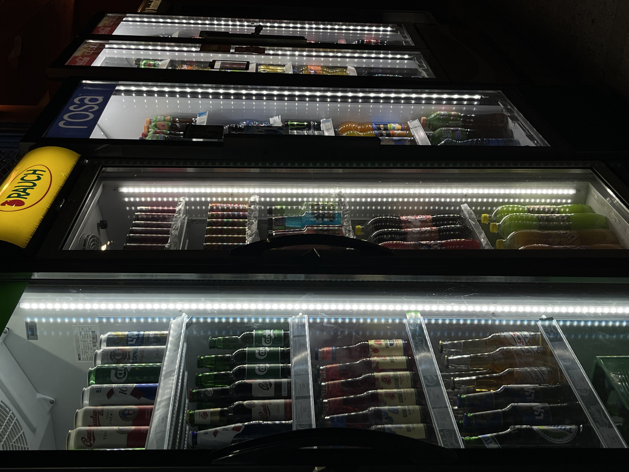 The image shows a row of commercial refrigerators filled with various beverages, located in Budva, Montenegro. The refrigerators are illuminated by LED strips, casting a bright light on the products. In the foreground, the left-most refrigerator displays cans of beer, followed by glass bottles of beer and ciders on lower shelves. The second refrigerator from the left contains a variety of bottled drinks, including some with colorful liquids. The third refrigerator, labeled "ROSA," holds bottles of water and other beverages. The subsequent refrigerators display more bottled drinks, including some large orange bottles. The overall environment appears to be an indoor commercial setting, possibly a convenience store or a bar, with a dark background. No people are visible in the image.