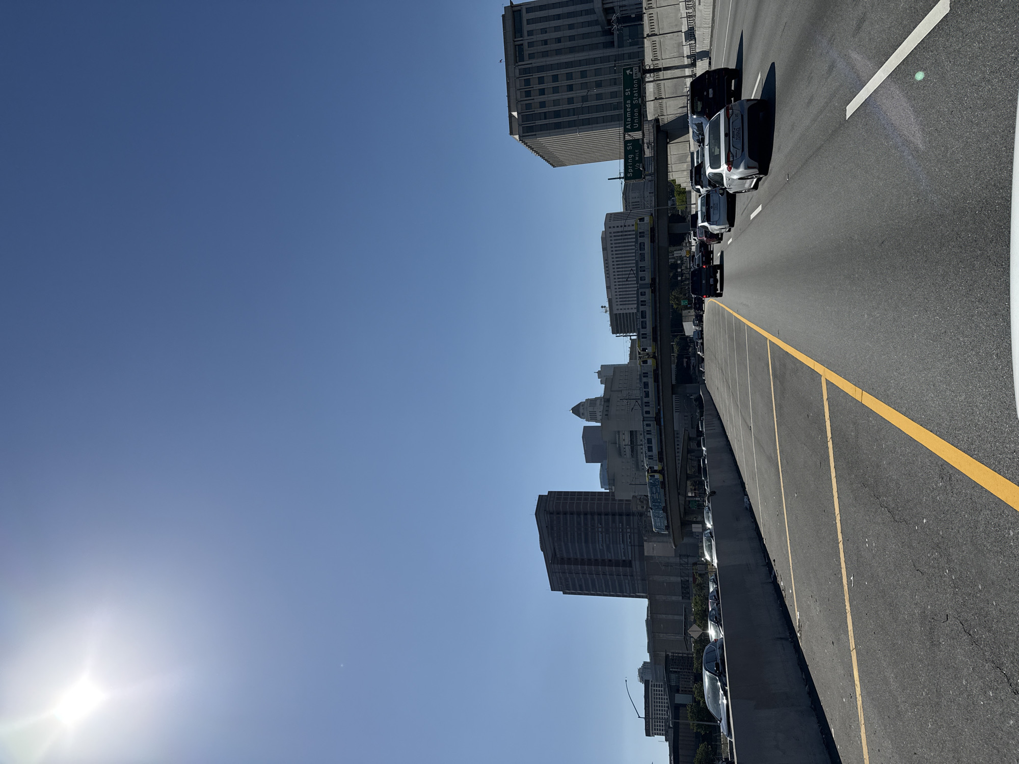The image depicts a view from a freeway in downtown Los Angeles, California. The sun is positioned low in the sky. Traffic is visible on the adjacent freeway lane. Downtown buildings line the freeway.