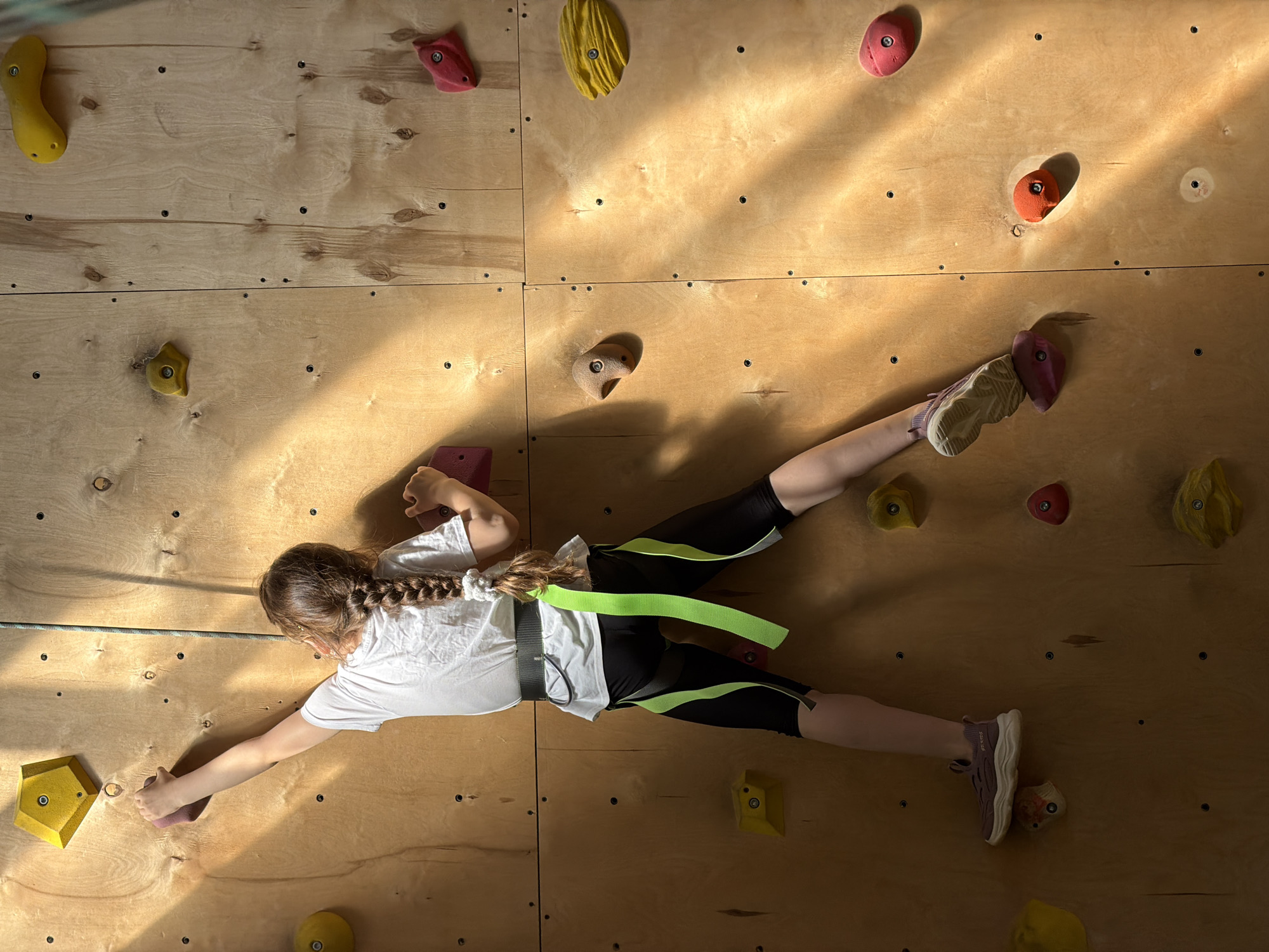 A girl is rock climbing on an indoor wall. She wears a white t-shirt, black leggings with neon green stripes, and climbing shoes. She is harnessed and connected to a climbing rope. The climbing wall is made of plywood and has various colored climbing holds attached to it. The location and city are not discernible from the image.