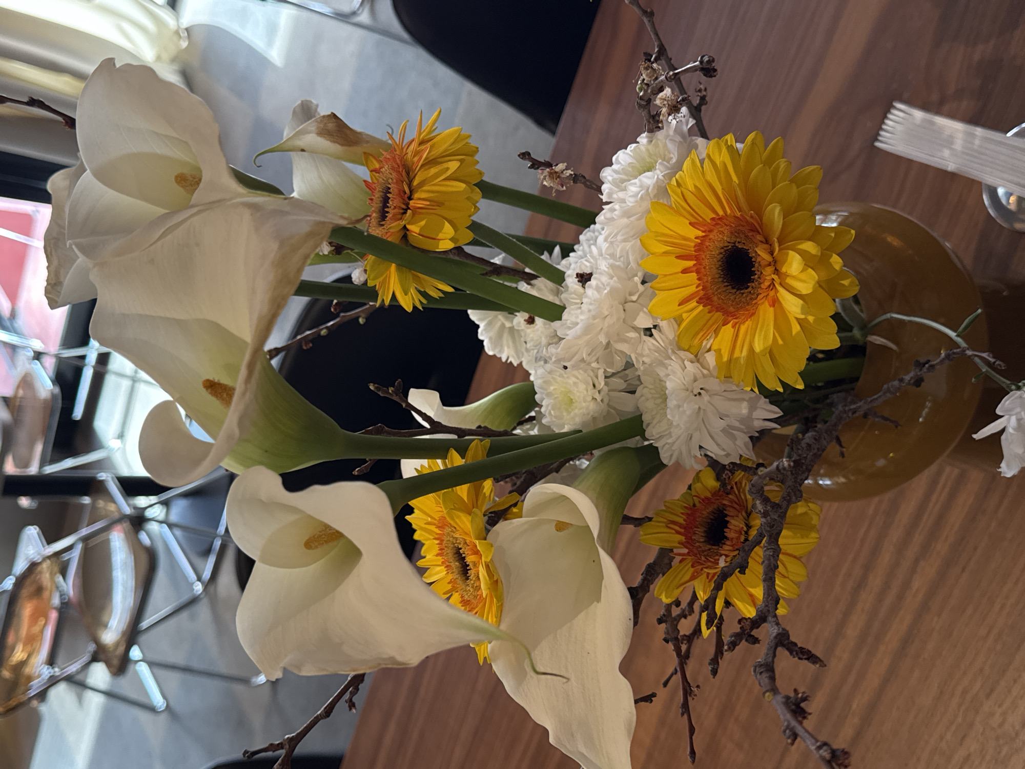 A still-life image captures a floral arrangement in an amber-colored vase set on a wooden surface. The arrangement consists of white calla lilies, yellow gerbera daisies, and smaller white flowers resembling chrysanthemums. Bare branches also extend from the arrangement, adding texture. The background includes a partially visible chair and what appears to be a window or glass partition, with some hints of the outside visible. Silverware is visible on the upper right, adding a dining aspect to the scene. The scene does not contain any people. The exact location in Yerevan, Armenia cannot be determined from this image.
