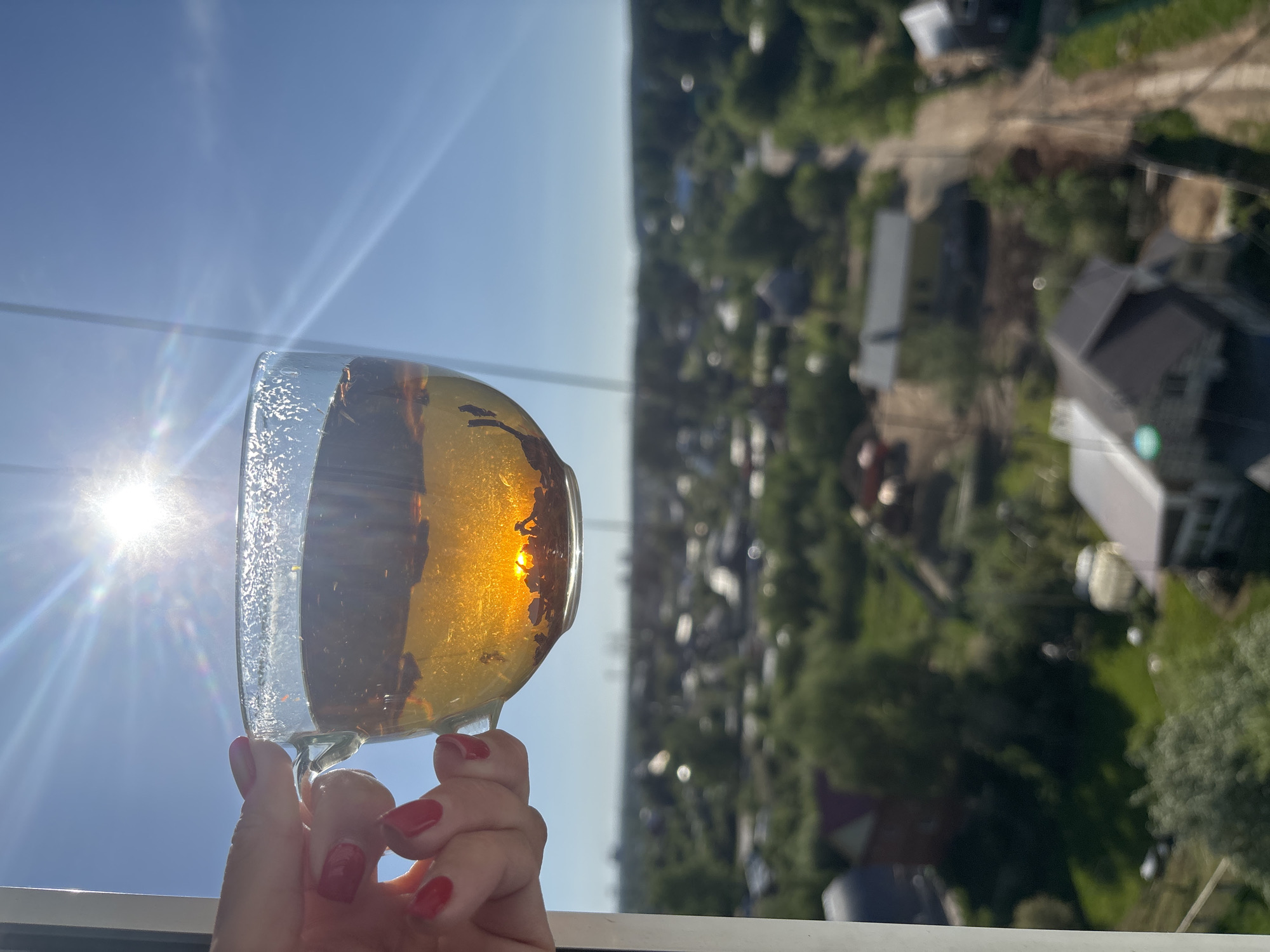 A hand holding a glass of tea in front of an out-of-focus cityscape under a sunny sky. The tea is amber colored with visible tea leaves. The sun shines brightly behind the glass. The hand has red nail polish. The cityscape background appears to be a residential area with green trees and rooftops. The photograph was taken in Chaikovsky, Russia.