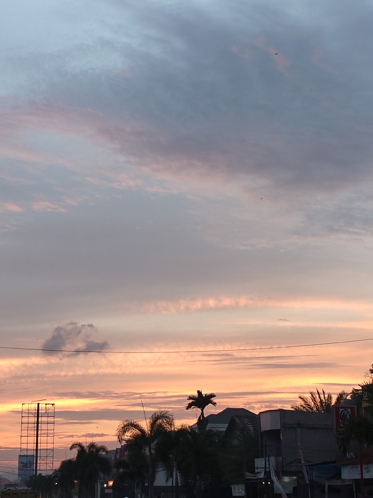 A serene dusk scene unfolds over Lhokseumawe, Indonesia, with the sky painted in soft hues of pink, orange, and blue. Wispy clouds stretch across the canvas, catching the last rays of the setting sun. Silhouetted against this vibrant backdrop are palm trees, their fronds reaching towards the sky, and the angular structures of buildings and a prominent billboard. The environment suggests an urban setting, possibly a roadside view, with hints of electrical wires crisscrossing the frame. In the distance, two small, dark objects are visible against the sky, possibly birds or kites, adding a touch of life to the tranquil atmosphere. The overall impression is one of peace and quiet beauty at the end of the day.