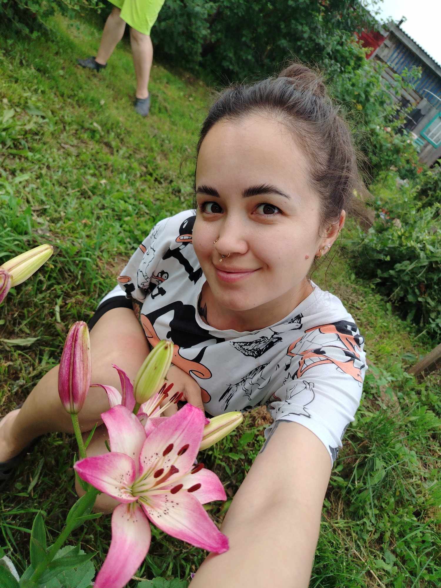 In the city of Gozhan, Russia, a young woman with dark hair tied in a bun, wearing a white t-shirt with a cartoon print, sits on a grassy area. She is holding a pink lily flower and its buds. A second person, wearing a bright green skirt and dark shoes, is visible in the background, partially obscured by foliage. The environment includes lush green grass, trees, and a rustic wooden structure with a blue and red roof, likely a house, in the distance. The lighting suggests an outdoor, daytime scene.