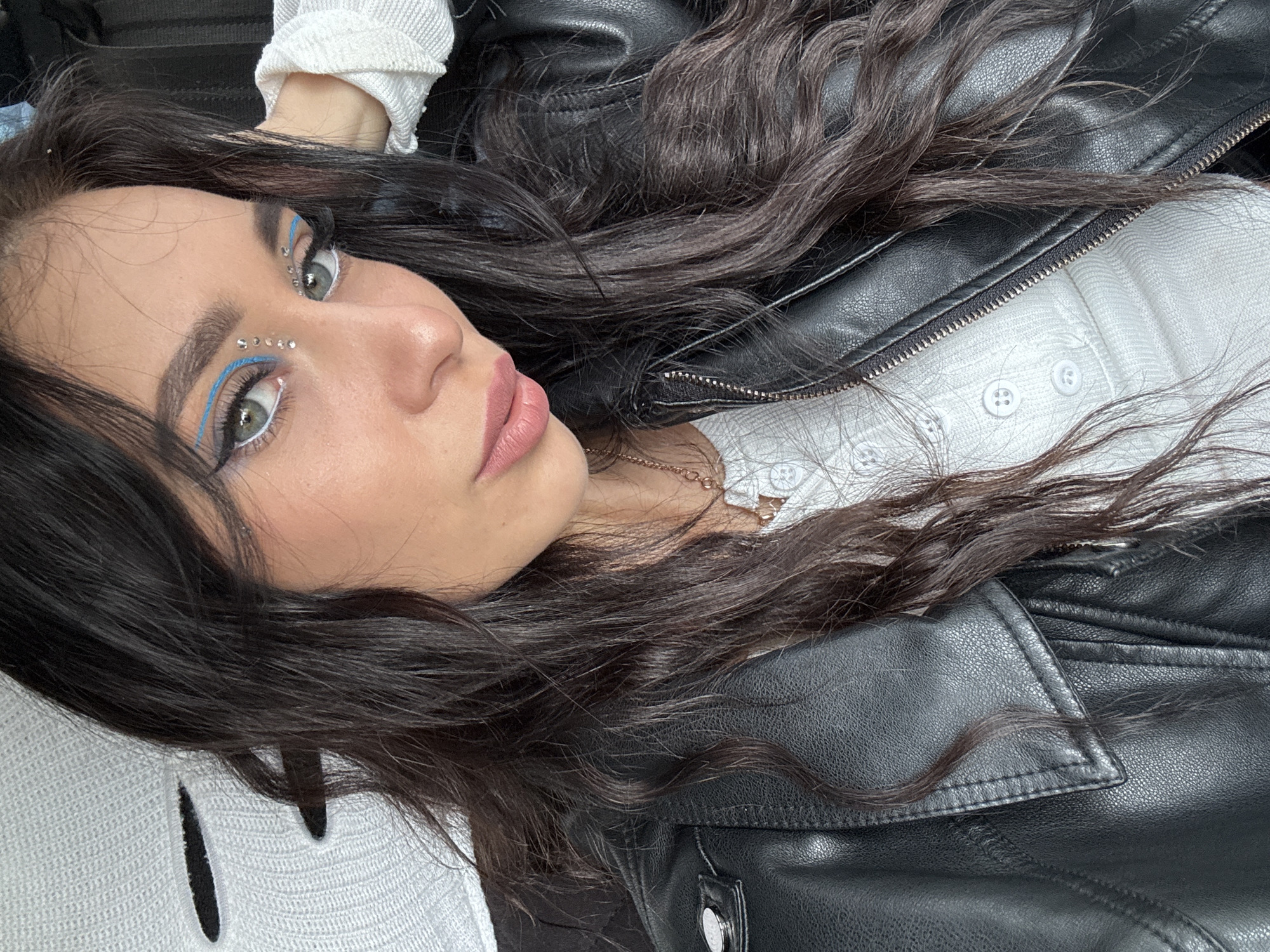 A brunette woman with blue eyeliner and small decorative gems applied to her face lies on her back, with her head angled towards the camera. She is wearing a white, button-down shirt under a black leather jacket. Her dark hair is spread around her head and shoulders, partially obscuring the shirt and jacket. The background is indistinct. The location is unknown and there is no discernible indication of Фоки, Russia.