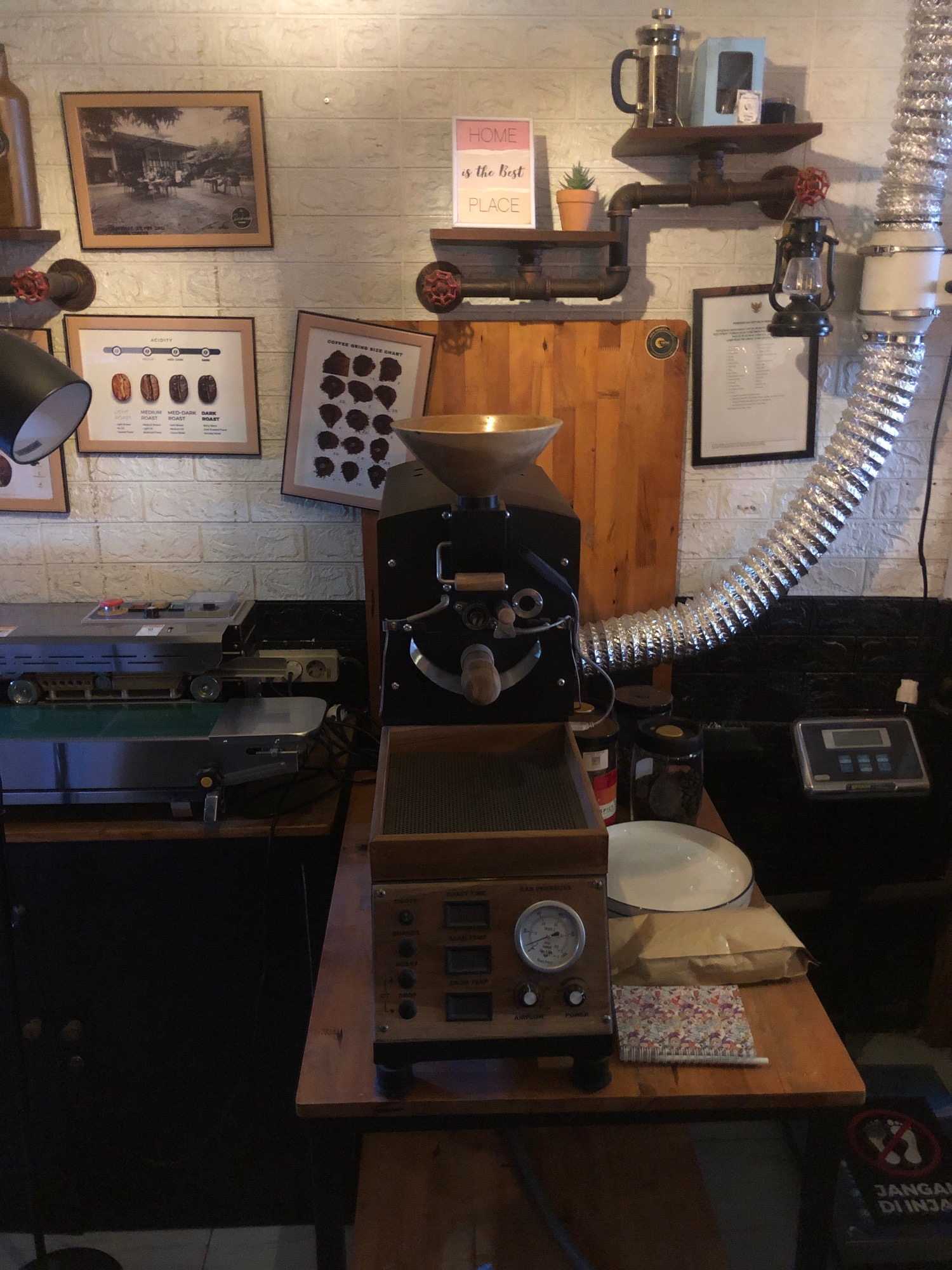 The image depicts the interior of a coffee shop, likely located in Lhokseumawe, Indonesia, given the sign in the lower right corner that reads "JANGAN DIINJAK" (Do Not Step). The scene is set against a white, textured brick wall. On a shelf above, a French press coffee maker, a blue box, and a small potted plant are visible. Below this shelf, a framed photograph of a group of people sitting at tables outdoors is displayed. To the right of this photo is a framed chart titled "COFFEE GRIND SIZE CHART" and another framed document.
In the foreground, a coffee roasting machine is prominently featured, with a metal funnel on top. To the left of the roaster, a sealing machine is visible. Various jars containing coffee beans, a white plate, and a small notebook with a floral pattern rest on a wooden table in front of the roasting machine. A digital scale is also present to the right of the roaster. The overall environment suggests a functional coffee preparation area, possibly with a rustic or industrial aesthetic, indicated by the use of metal pipes and exposed machinery.