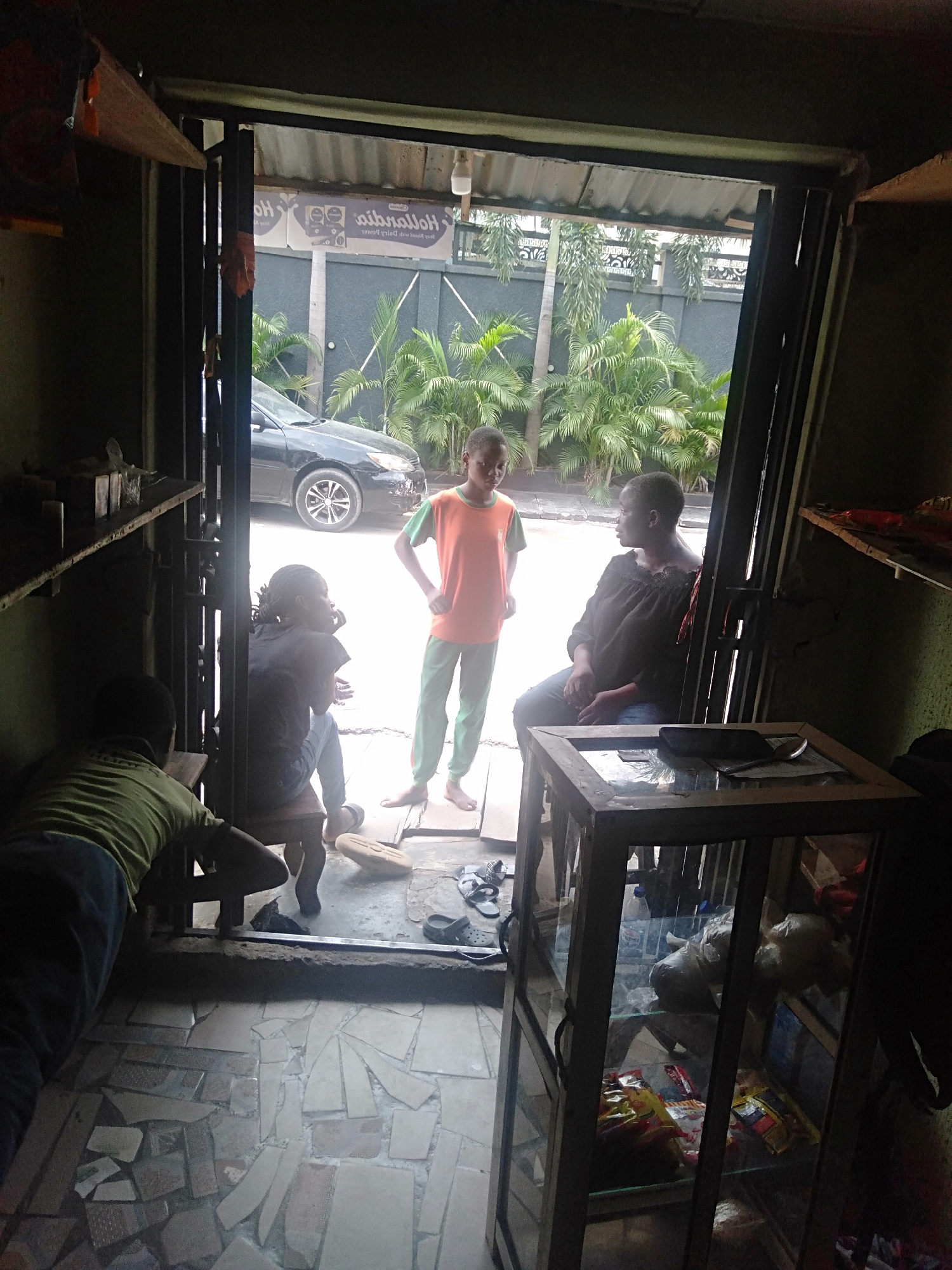 The image captures an indoor perspective looking out through a doorway into a bright outdoor scene. The interior is dimly lit, revealing a small shop or room with dark wooden shelving along the walls. On the right, a tall, slender wooden display cabinet with glass panels contains various packaged goods and white plastic bags; a mobile phone and spoon rest on its top surface. The floor is made of irregularly shaped, broken tiles.
Four individuals are visible. Inside, on the far left, a person (likely a child or young adult) in a green t-shirt and dark pants is hunched over a low wooden bench, facing away from the camera. At the threshold of the doorway, a second person (child or young adult) in a black top is seated on a low stool, facing inwards to the left. Outside the doorway, a young person in an orange t-shirt with green sleeves and green pants stands barefoot with hands on hips, looking towards the two seated individuals. Further inside on the right, an adult female in a dark, off-shoulder top and jeans sits on a low stool, looking left, seemingly at the group near the entrance.
The exterior shows a bright daytime setting with a dark sedan parked on a paved surface. Behind the car, a grey concrete fence with decorative elements is flanked by lush green tropical plants, including palm trees. A corrugated metal roof overhangs the doorway. Above the car, a sign is visible but inverted and backwards, displaying "Hollanbrdia" (likely "Hollandia" reversed) and "Quality Drinking Yogurt from FrieslandCampina," along with a partial "Ho." The scene depicts a casual gathering or conversation at the entrance of what appears to be a small business, likely in a warm climate.