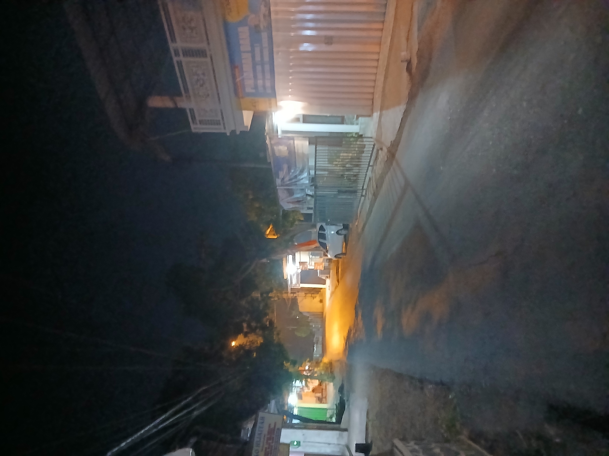 A nighttime street scene in Kademangan Kulon, Indonesia, depicts a road with some illuminated buildings and a parked white car. On the left side of the road, a building with a sign that reads "AO HIJAYAH" is visible. Further down the street, there are more buildings with lights on. To the right of the road, a building with a metal gate and a white corrugated metal facade is prominent, with a sign above it that appears to say "PINJAMAN DANA". A white car is parked on the right side of the road, facing away from the camera. The street is asphalt and appears to be in a somewhat rural or suburban area, with trees and power lines also present. The lighting suggests it is dusk or night, with artificial lights from the buildings illuminating the scene.