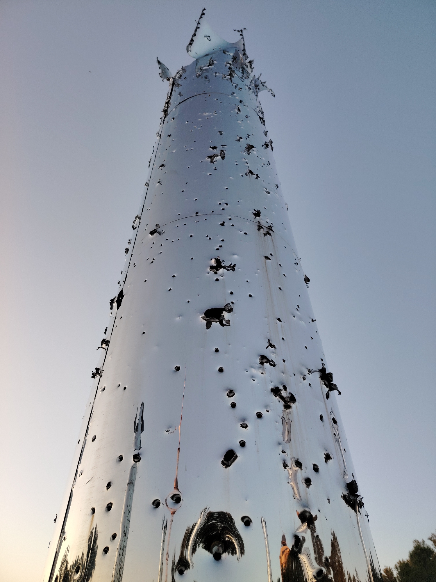 The photograph captures a low-angle view of a tall, cylindrical, reflective monument in Kyiv, Ukraine. The monument appears to be made of polished metal and is heavily damaged, with numerous holes and tears across its surface, particularly concentrated towards the top, which is jagged and appears to be partially destroyed. The surface reflects the sky, which is a pale, clear blue, and hints of trees at the bottom of the frame suggest an outdoor setting. The damage to the monument strongly implies it has been subjected to impact, possibly from shelling or explosions, given the context of the city. There are no people visible in the frame. The monument's distressed state is the primary focus, conveying a sense of destruction within an urban or park-like environment.