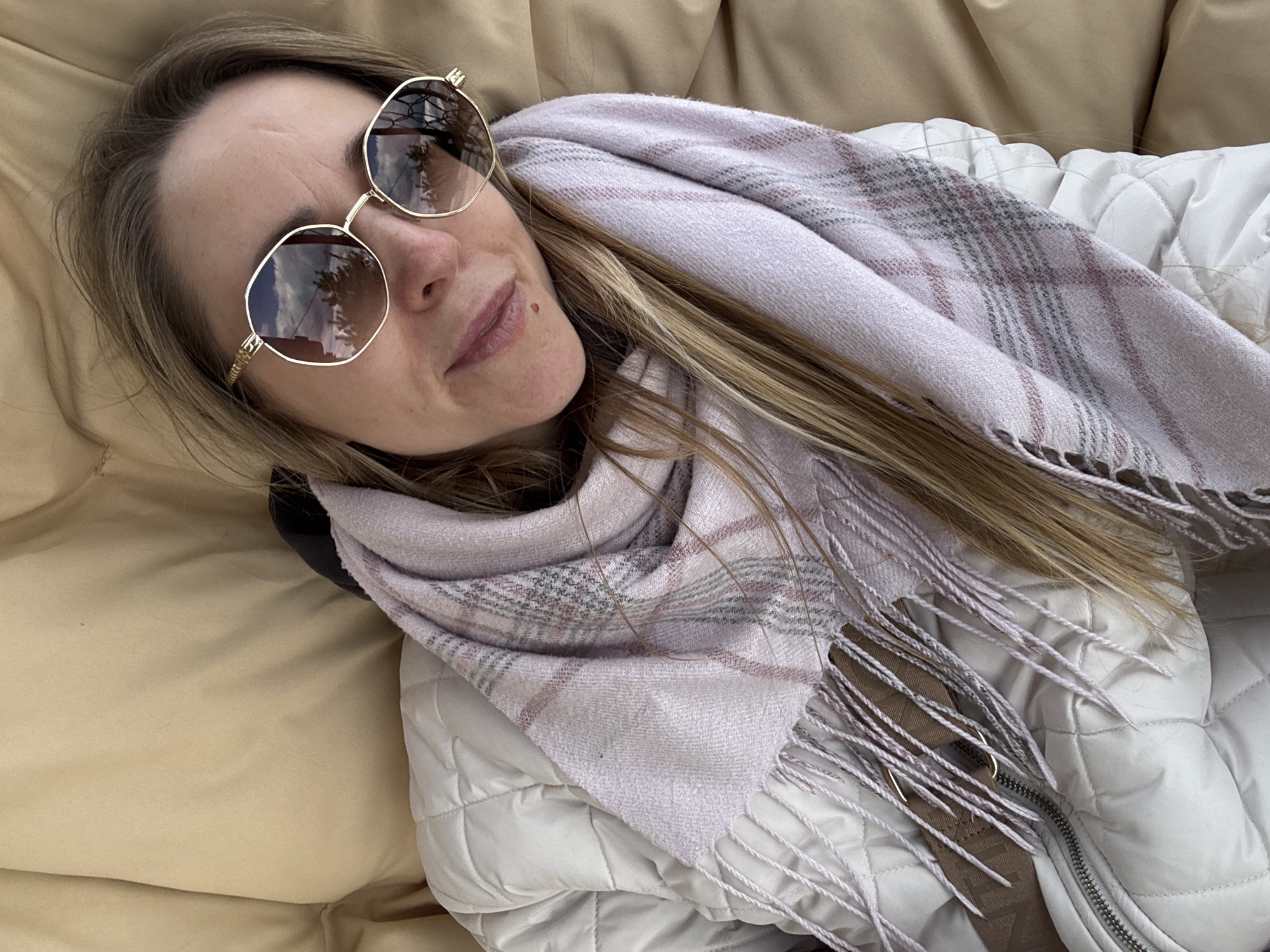 The image features a woman reclining, captured from a slightly overhead perspective, in what appears to be a relaxed setting in Chaikovsky, Russia. She has long, light brown or blonde hair and is wearing a light-colored, possibly off-white, quilted jacket and a soft, light pink or lavender plaid scarf with fringed edges.
The woman is wearing large, round sunglasses with gold-colored frames and highly reflective lenses. The reflections in her sunglasses clearly show a bright, cloudy sky and the silhouettes of trees, suggesting an outdoor, daytime scene. Her facial expression conveys a sense of contentment, with a slight, relaxed smile. A small mole is visible on her chin.
The background consists primarily of a plain, soft tan or beige fabric, likely a cushion, blanket, or part of a lounge chair. The quilted texture of her jacket and the scarf suggest cooler weather conditions. Faint, indistinct dark text, possibly "ANZA," is partially visible on a brown strap located near the zipper of her jacket on the right side of the frame.