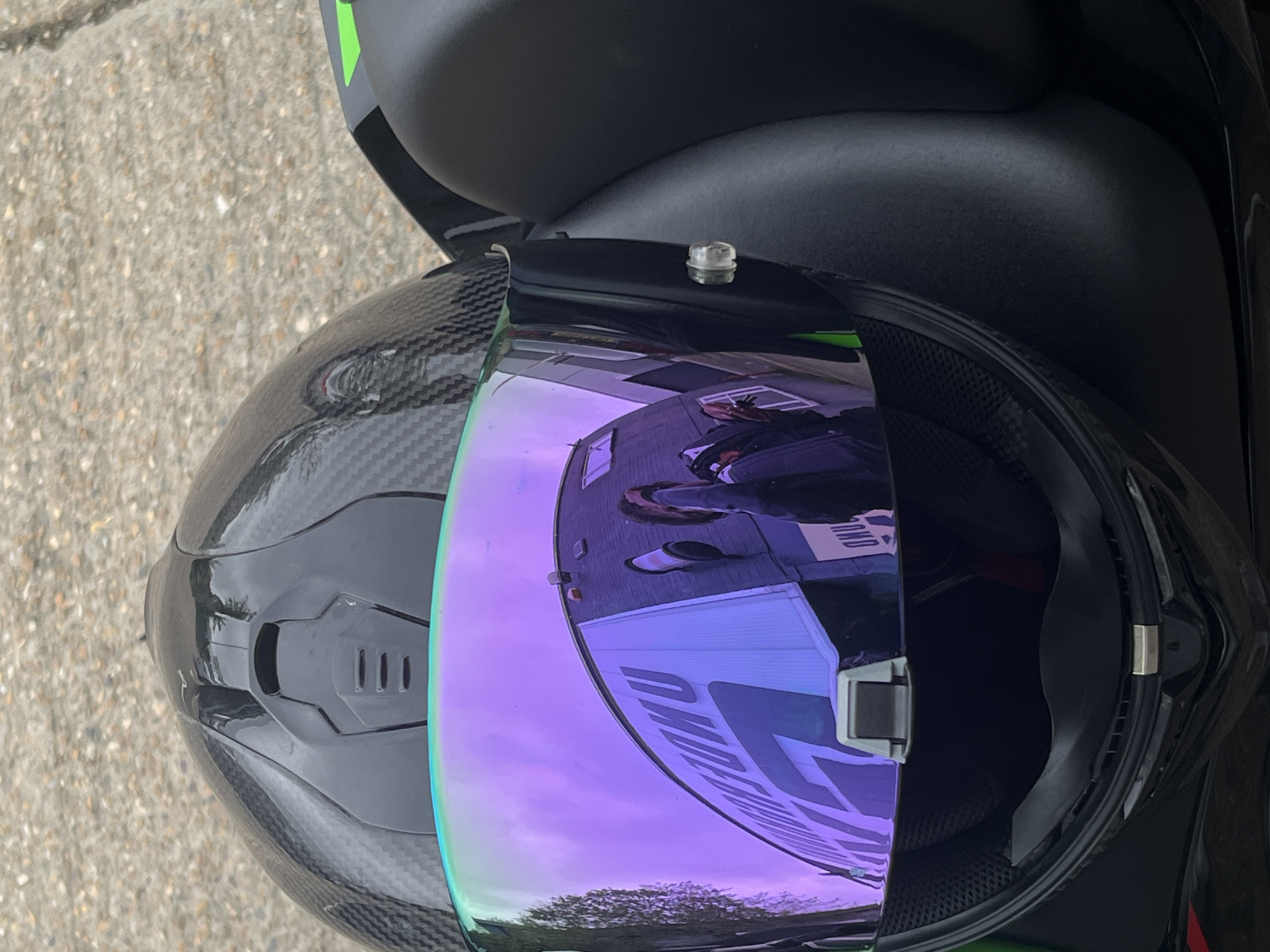 A top-down shot displays a black motorcycle helmet, presumably resting on the motorcycle's seat. The helmet is mostly black, and the visor has a purple/blue tint, mirroring the surrounding environment, revealing a partial reflection of a building, signage, a person, and trees. The pavement is visible in the background. The photograph appears to be taken in Tunbridge Wells, United Kingdom, as the reflection within the visor indicates a building with lettering that potentially says "Tunbridge".