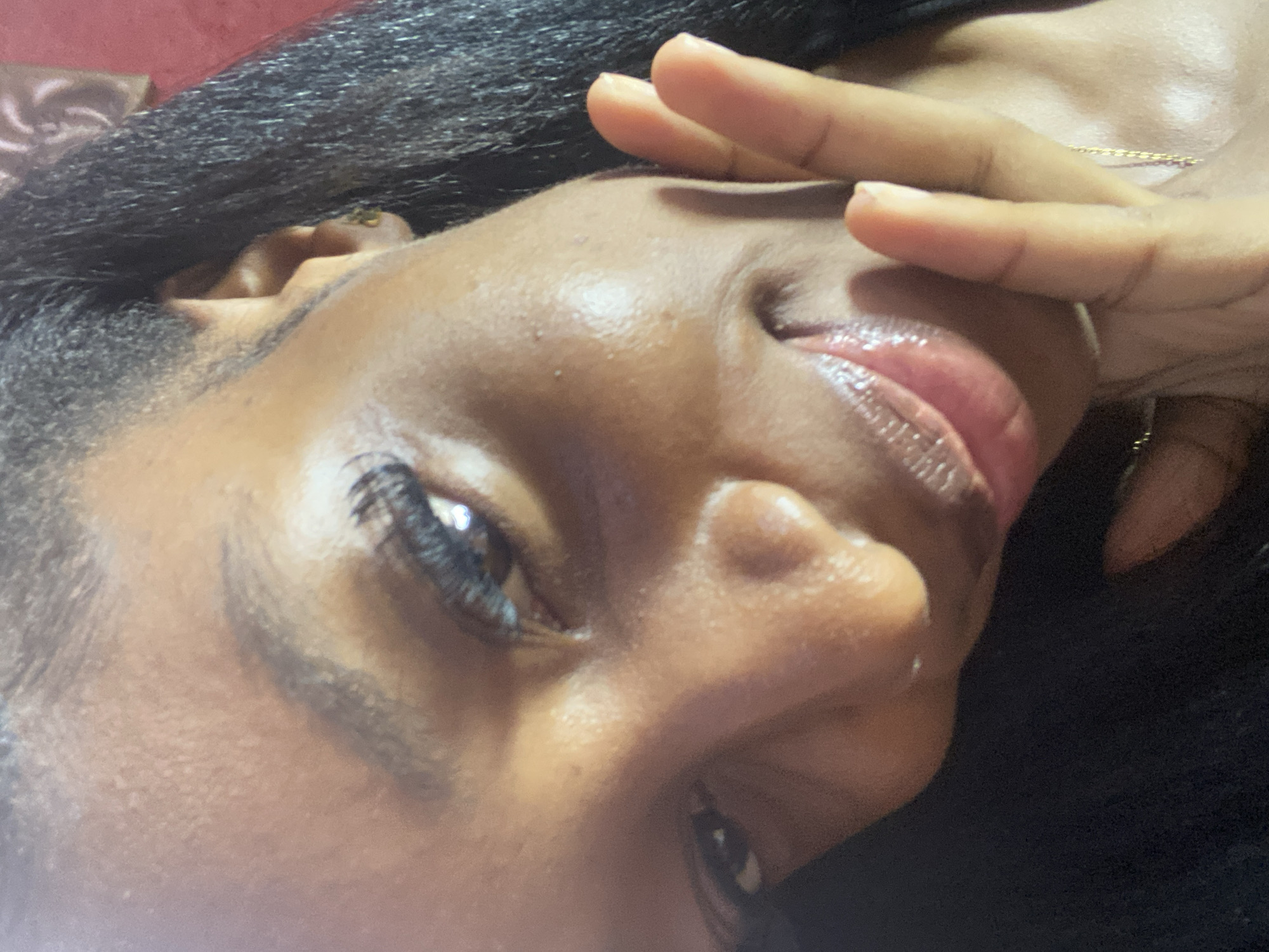 This is a close-up, high-angle, selfie-style shot of a young Black woman in Jos, Nigeria. She is positioned in the lower half of the frame, with her head tilted and her face partially obscured by her hand. Her left eye is visible, with long eyelashes and dark makeup. Her lips are glossy and parted. A delicate gold chain is visible around her neck. The background is out of focus, showing a reddish-brown textured surface and dark hair. The lighting is bright and appears to be coming from above and to the right.
