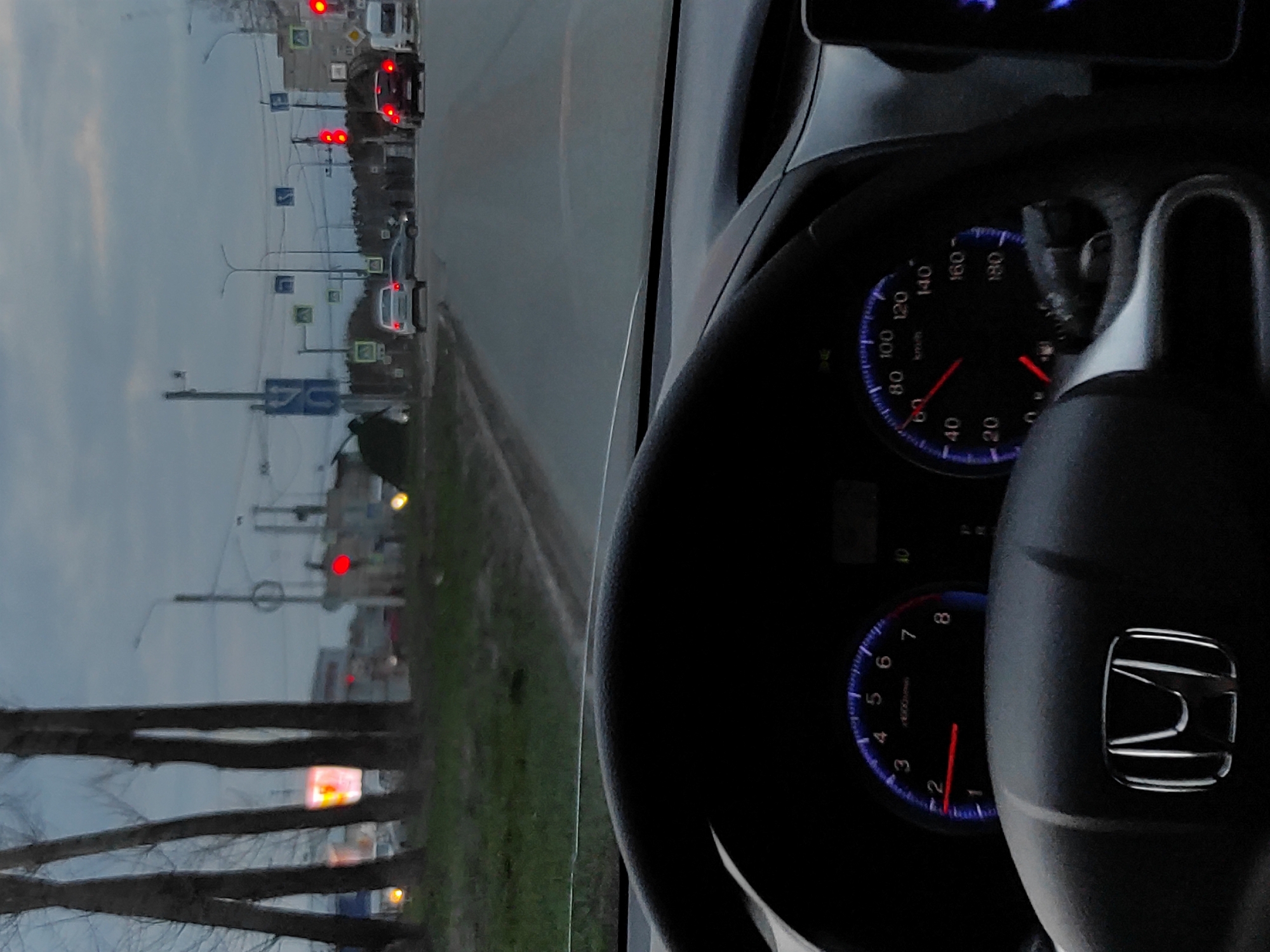 The image captures a driver's view from inside a stationary car, looking out onto a road in Chaikovsky, Russia, during what appears to be dusk or early evening under an overcast sky.
In the immediate foreground, a black steering wheel with a prominent silver Honda logo occupies the lower right side. To the left of the steering wheel, the car's dashboard is visible, featuring illuminated gauges with blue backlighting. The speedometer clearly shows a reading of 0 km/h, while the tachometer also indicates 0 RPM, confirming the vehicle is stopped. The text "km/h" is visible on the speedometer.
Beyond the windshield, a multi-lane asphalt road stretches into the distance. Several cars are stopped ahead, with their red brake lights illuminated, indicating they are waiting at a traffic light. Two distinct red traffic lights are visible, signaling a stop for the vehicles. Alongside the road, various road signs are posted on poles, including a blue square sign with a white arrow pointing straight, another blue sign with the number "70" (likely an informational sign), and several diamond-shaped signs, characteristic of 'Main Road' indicators in Russia. Utility poles with overhead wires line the roadside. Bare trees, suggesting a colder season like late autumn or early spring, are prominent on the left side of the frame. The background is indistinct, showing a grey, hazy sky and faint outlines of distant buildings. The overall scene depicts a common urban traffic situation at the close of day.