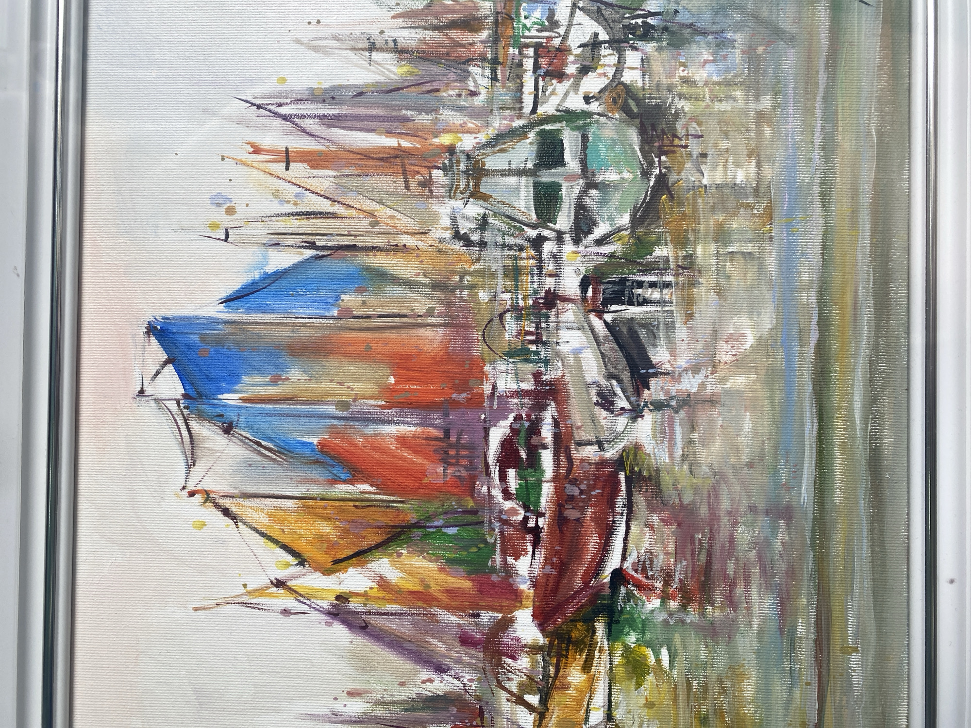 The image shows a painting of a scene in Varna, Bulgaria. The painting is framed. The left side depicts colorful sailboats, possibly in a marina. The right side suggests a street scene with buildings and vertical lines. The painting style is abstract and expressive, with visible brushstrokes and a mix of colors. There are no people visible in the painting.