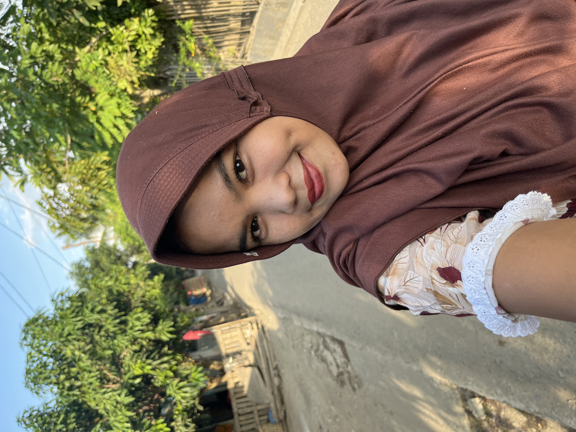 In Kruengmate, Indonesia, a young woman with a brown hijab and a floral patterned shirt takes a selfie on a paved road. The woman has dimples and is wearing red lipstick. Sunlight casts shadows on the road and the surrounding trees and buildings. A white lace trim is visible on her sleeve. The background depicts a rural village scene with houses and vegetation.