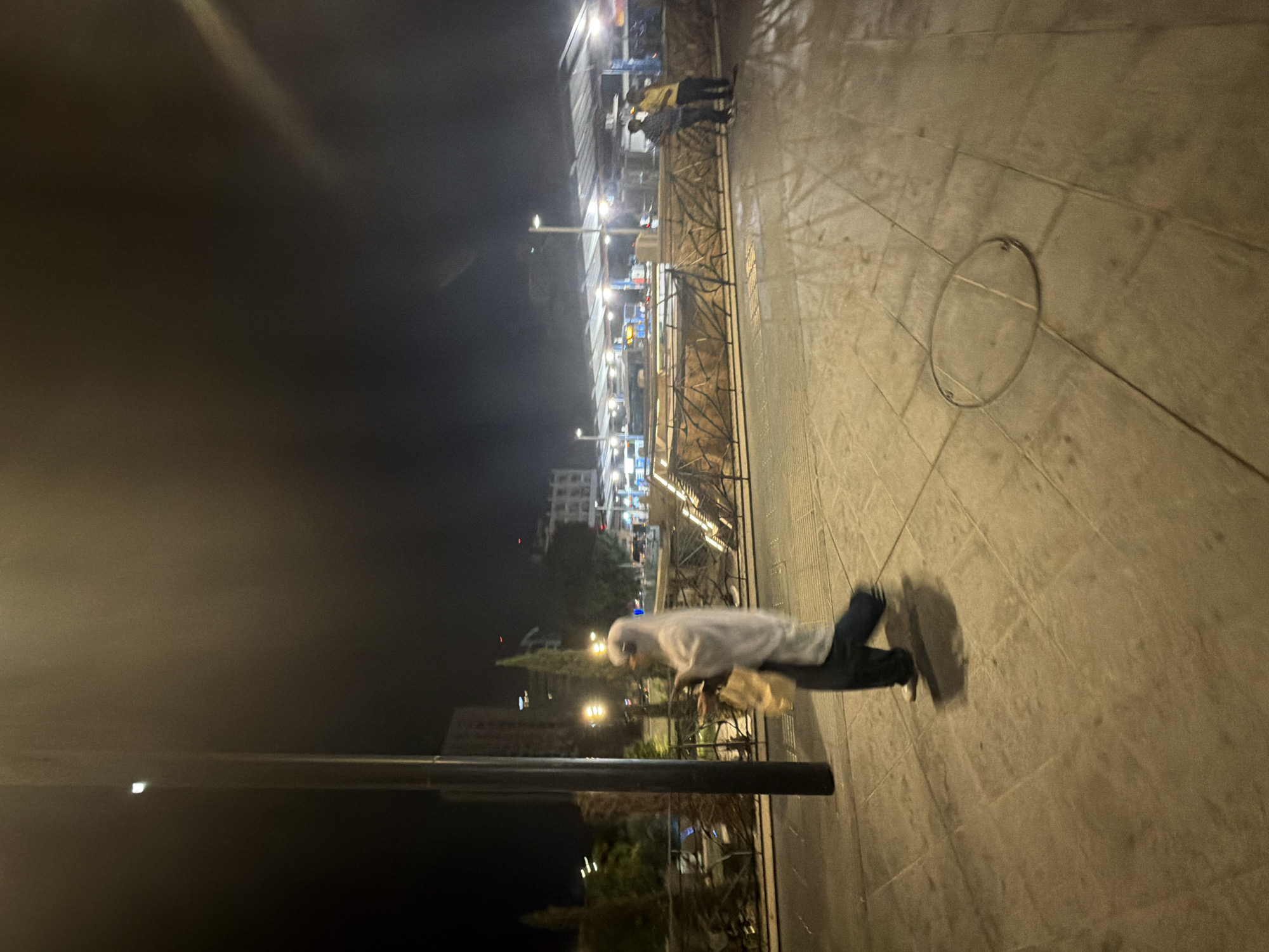 The image captures a nighttime scene in Nicosia, Cyprus. A person is walking on a paved area, appearing to be a public space like a promenade or sidewalk. The person is wearing a white hooded jacket and dark pants, and is carrying a shopping bag. They appear to be walking away from the camera.
In the background, there are several buildings with lights on, suggesting a commercial or residential area. There's also a fence with an ornate design, and streetlights illuminating the walkway. The overall atmosphere is dimly lit, typical of a night scene. The paving stones are clearly visible, and there is a circular metal fixture embedded in the ground. The image has a slightly blurred effect, possibly due to low light conditions or movement. There is no visible text in the image.