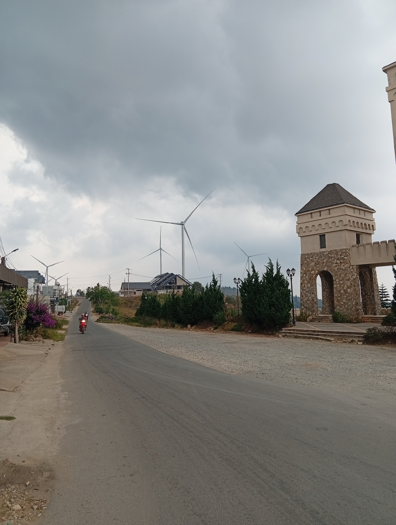 This image captures a daytime scene along a road under a heavily overcast and cloudy sky. The perspective is from the roadside, looking down a gently sloping asphalt road that recedes into the distance.
On the left side of the road, there is a concrete sidewalk bordering residential buildings and what appears to be a small commercial establishment or home. Lush green and purple flowering plants grow along this side. A red motorcycle with two helmeted riders is visible on the road, heading away from the viewer. Further down the road on the left, several modern buildings with dark, angled roofs (possibly incorporating solar panels or unique architectural styles) are visible, along with utility poles and power lines.
The most prominent features in the background and to the right are several large wind turbines with white blades and towers, indicating a wind farm. One turbine is particularly central, its blades contrasting against the gray sky.
On the right side of the foreground and mid-ground, there is a distinctive, castle-like stone structure. This building features arched gateways and a square turreted tower, constructed with stone blocks at the base and a lighter-colored upper section with crenellations and small windows. It appears to be an entrance or a decorative element to a property or attraction, complete with steps leading up to it. Various green bushes and small trees are neatly planted around this structure and along the right-hand dirt/gravel shoulder of the road. Streetlights are also visible near the castle-like building.
The overall scene combines elements of modern renewable energy infrastructure, residential living, and unique, themed architecture within what appears to be a semi-rural or developing area. The cloudy sky suggests a muted, soft light throughout the scene. No discernible text is visible.