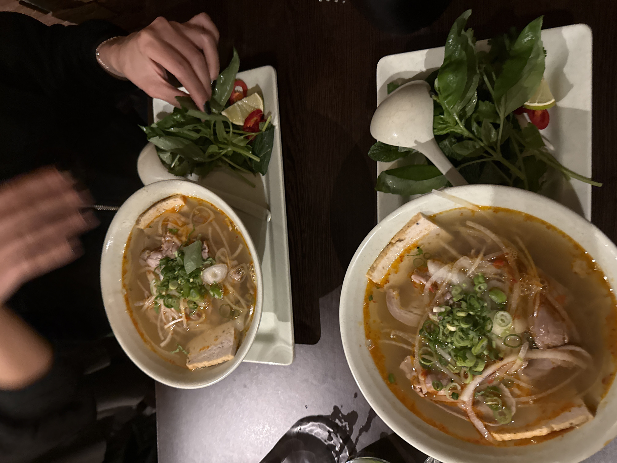 The image shows two bowls of Vietnamese noodle soup, Bun Bo Hue, on a dark table, possibly in a restaurant in London, United Kingdom. A person is adding herbs from a side plate to the left bowl. The side plates also contain lime and chili. Each bowl has a spoon. The lighting is dim.