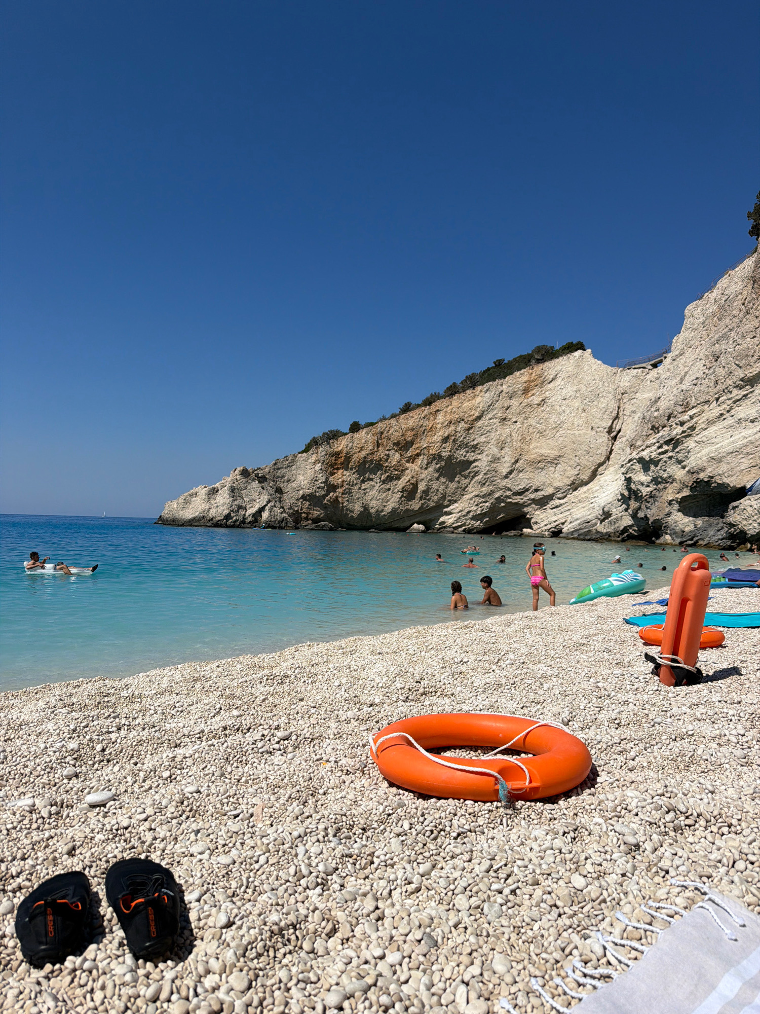 A bright, sunny day is depicted at a pebble beach in the Municipal Unit of Apollonioi, Greece. The clear, turquoise water of the Ionian Sea laps gently onto the shore. A person is relaxing on an inflatable raft in the water. Several people are swimming and playing in the sea.
On the beach, a young girl wearing a pink swimsuit and goggles stands near a green inflatable toy. An orange life preserver ring and a larger orange flotation device with rope are visible on the pebbles. In the foreground, a pair of black water shoes with orange accents rests on the beach. A light-colored beach towel with white stripes lies to the right.
Towering limestone cliffs, covered in sparse greenery, form a dramatic backdrop to the scene. The sky is a deep, cloudless blue. The overall impression is one of a serene and idyllic summer day at a popular coastal destination.