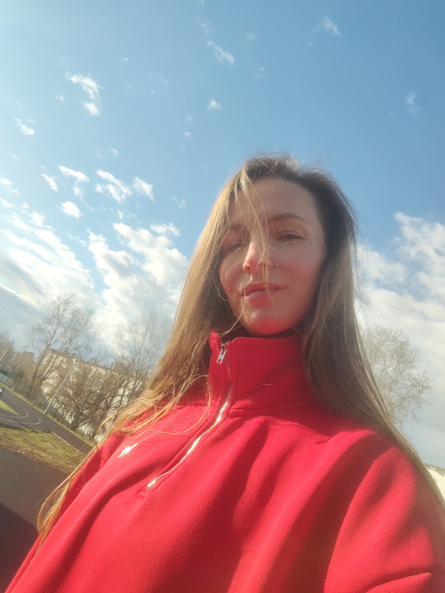 This is a low-angle outdoor selfie featuring a woman with long, light brown hair, captured on a bright, sunny day in what appears to be Chaikovsky, Russia. She is positioned in the lower-middle of the frame, looking directly upwards towards the camera with a subtle, content smile. Her hair is casually styled, with some strands falling across her face.
She wears a vibrant red fleece jacket or sweatshirt with a silver-colored half-zip at the neck. A small, indistinct white logo is visible on the upper left chest of her garment. The upper portion of the image showcases a vast, clear blue sky dotted with scattered white cumulus clouds.
The background beneath the sky reveals a landscape consistent with late winter or early spring, characterized by numerous bare trees. In the distance, several buildings can be vaguely seen. To the left, a portion of a dark asphalt running track or sports field is visible, bordered by some green grass, suggesting a recreational or park area. The strong sunlight casts bright highlights on her hair and face. No discernible text is visible in the image.