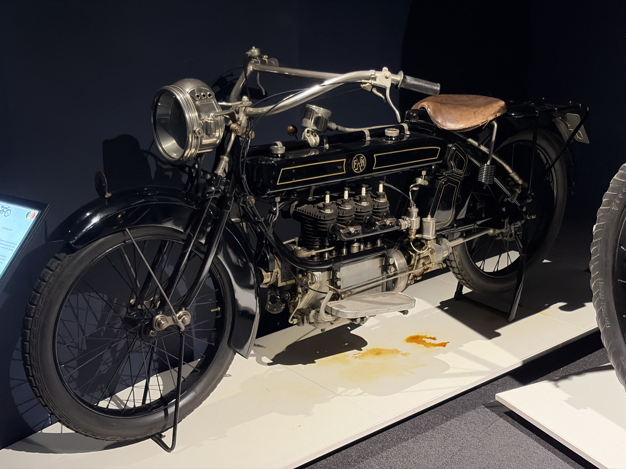 A vintage black motorcycle is centrally displayed in what appears to be a museum or exhibition setting. The motorcycle is predominantly black with gold pinstriping and a distinct "FN" logo on its fuel tank. It features a shiny chrome front headlight, exposed engine components including multiple spark plugs on an inline-four cylinder engine, and polished metal accents. A brown, well-worn leather sprung saddle sits atop the rear. The handlebars are also chrome, with classic levers and grips.
The motorcycle is stationary, presented on a bright white display platform. Beneath the engine area, a prominent brownish-orange stain, resembling spilled oil or coolant, is visible on the white platform, adding a touch of historical realism to the exhibit. The immediate background is dark, suggesting subdued lighting or spotlights focusing on the motorcycle.
On the far left, a glowing informational plaque is partially visible, though its text is unreadable. Another tire, partially cropped on the right, also rests on a white platform. Faint text on its sidewall indicates "FIRESTONE". The overall scene highlights the historical significance and engineering of the vintage vehicle.
