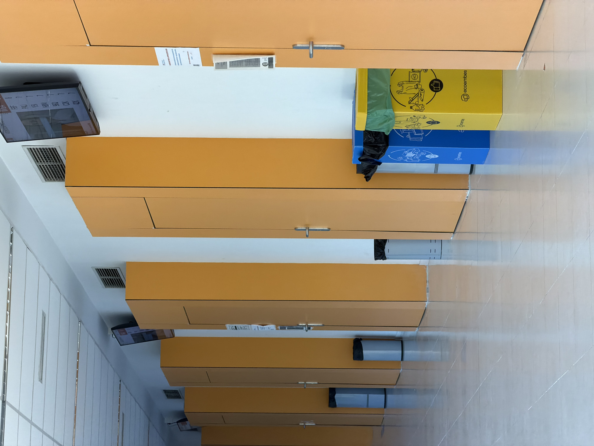 The image depicts a well-lit interior corridor with a polished floor that reflects the surroundings. The corridor is lined on the right with a series of tall, orange-colored doors, punctuated by the white wall between them. The doors have silver handles and some have visible vents or notices attached. On the left side of the corridor, there are similarly colored orange panels, interspersed with silver trash receptacles of various designs, some cylindrical and others rectangular with different colored bags. One rectangular receptacle in the foreground is yellow with the "ecoembes" logo and imagery related to recycling. Another is blue with similar imagery. Further down the corridor, more doors and trash receptacles are visible. Mounted on the wall above the orange panels on the left is a flat-screen television displaying what appears to be a queue or waiting list with alphanumeric codes and numbers. The ceiling is white and composed of acoustic tiles with linear lighting fixtures. The scene appears to be a public building, possibly an office or service center, given the presence of the waiting list display and recycling bins. The overall impression is one of neatness and order within a modern, functional space. Given the "ecoembes" logo, which is a Spanish organization focused on recycling, and the general style of the building, this location could be within Las Rozas de Madrid, Spain, where such public facilities are common.