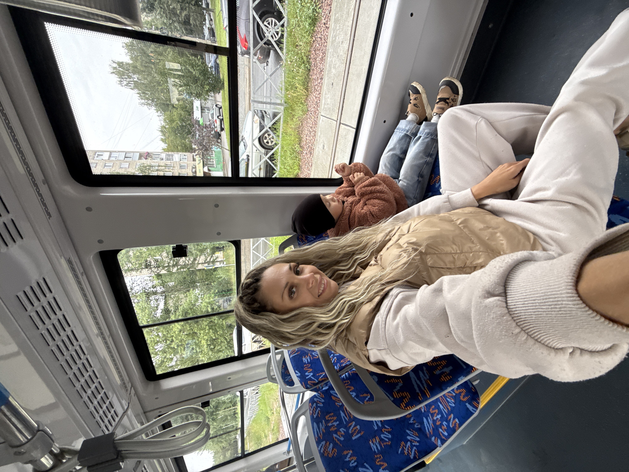 In Rusanovo, Russia, a woman and a child are seated inside a tram, looking out the window. The woman, with blonde hair styled in braids, wears a light-colored tracksuit and a tan puffer vest. She is positioned in the foreground, taking a selfie. Beside her, a child wearing a brown fuzzy jacket and a black beanie sits looking towards the window. The tram interior features blue seats with an orange and white pattern. Outside the window, a residential building and trees are visible, along with a street with cars and a tram track. The scene appears to be a daytime journey.