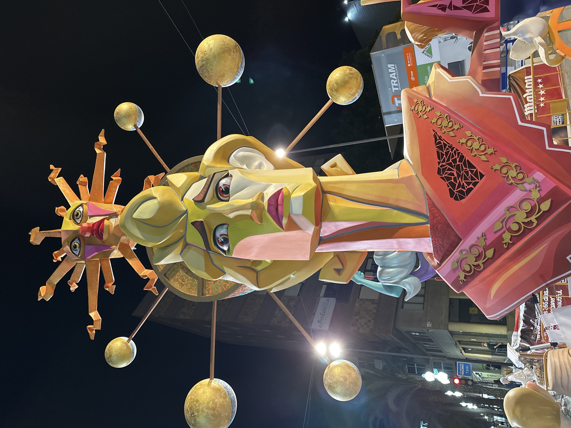 The image depicts a night scene in Alicante, Spain, featuring a large, colorful monument, likely a Falla, common during the Las Fallas festival. The monument is an elaborate sculpture of a face with a sun-like headdress. The face is geometrically constructed with vibrant colors, including yellow, pink, green, and red. The headdress consists of an orange sun with pointed rays and a separate face with similar features. Surrounding the central figure are several golden orbs, possibly representing planets or celestial bodies, suspended on thin rods.
In the background, buildings with windows are visible, some illuminated, suggesting an urban setting. Streetlights and traffic signals add to the nighttime ambiance. There are several palm trees lining the street. A prominent sign for "TRAM METROPOLITÀ D'ALACANT" is visible on a vehicle or structure in the lower right portion of the image, confirming the location as Alicante.
Several people are present, though their details are indistinct due to the low light and distance. Some appear to be walking along the street, while others might be observing the monument. Signs for businesses like "MERLIN" and "Mahou" are also visible, further contextualizing the urban environment. The overall scene captures a festive atmosphere with the impressive monument as the focal point.