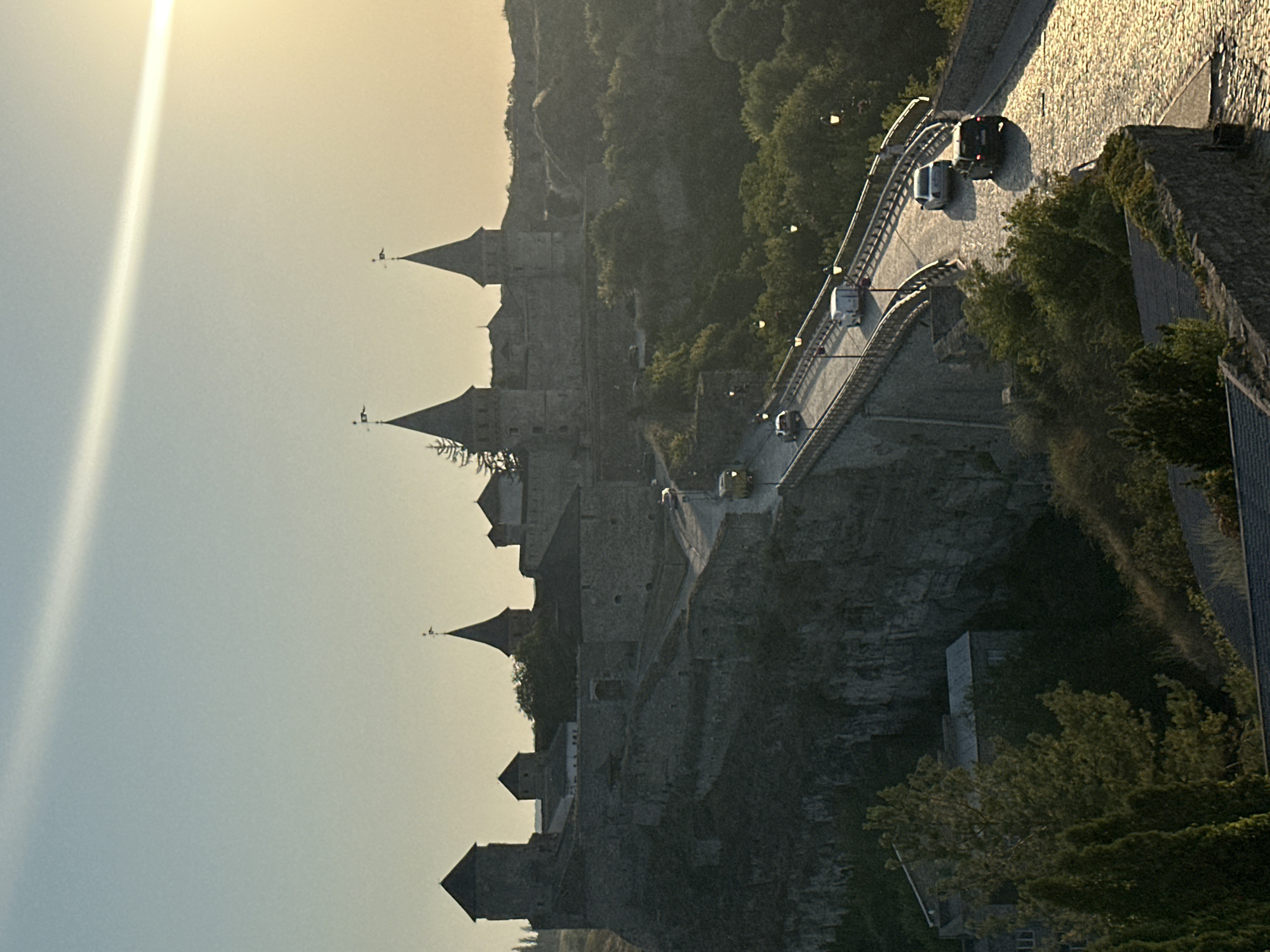 This image depicts the Kamianets-Podilskyi Castle in Kamianets-Podilskyi, Ukraine, during what appears to be late afternoon or early evening, with the sun casting a warm glow and creating long shadows. The castle, a formidable stone fortress with multiple towers, is situated on a rocky promontory. A paved road, lined with lampposts, leads across a bridge to the castle. Several vehicles, including cars and a yellow bus, are visible on the road and bridge. The surrounding landscape is characterized by lush greenery and rocky cliffs. The sky is a soft blue with a streak of sunlight extending across it.