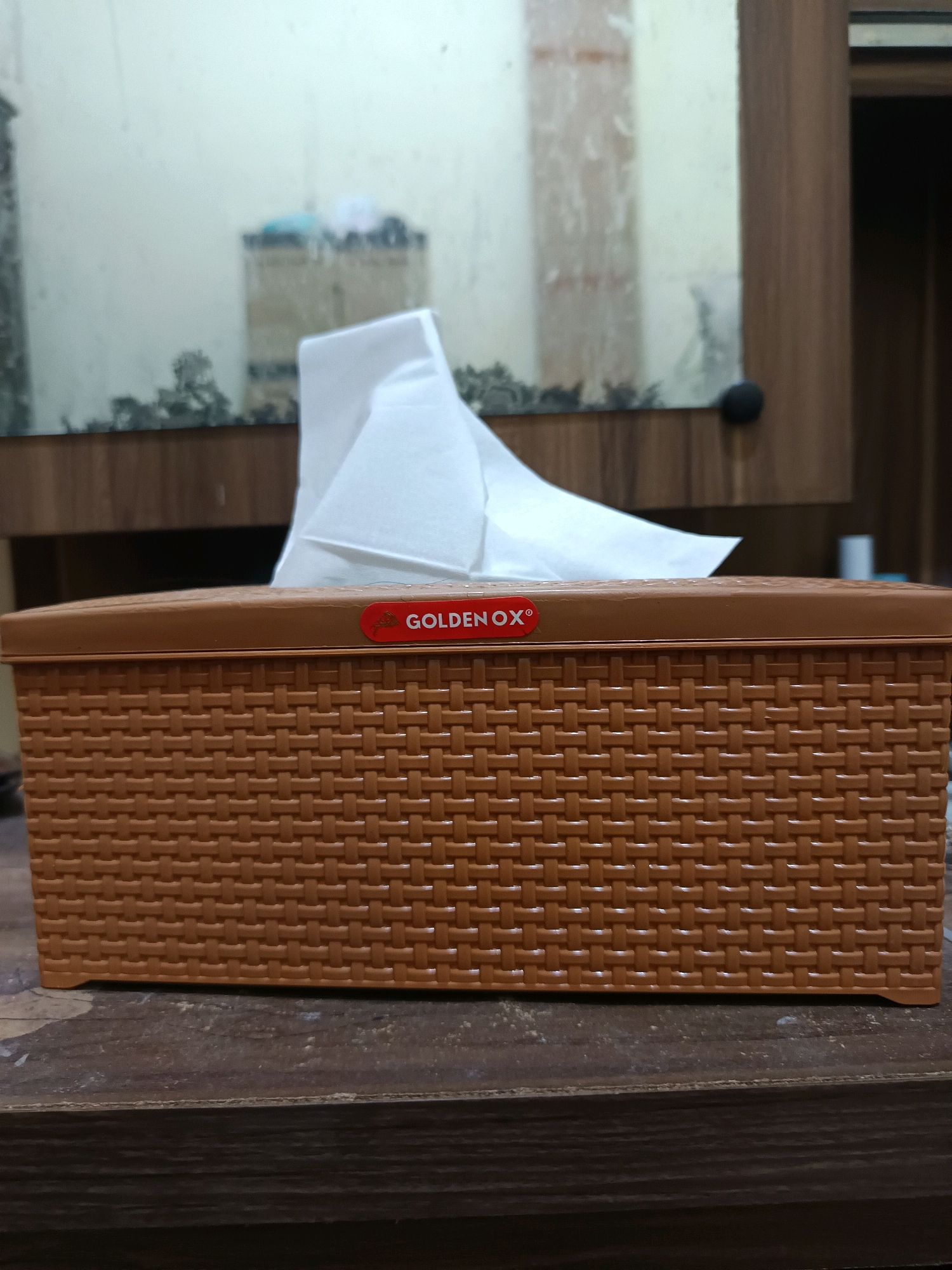 The image shows a close-up of a brown woven tissue box with a red sticker that says "GOLDEN OX". White tissues are pulled out of the top of the box. The tissue box is placed on a dark wooden surface. In the background, there is a mirror reflecting a pale wall and some dark objects. The scene appears to be indoors, possibly on a table in a room in Gunung Sekar, Indonesia, though the specific location details are not visible in the image itself.