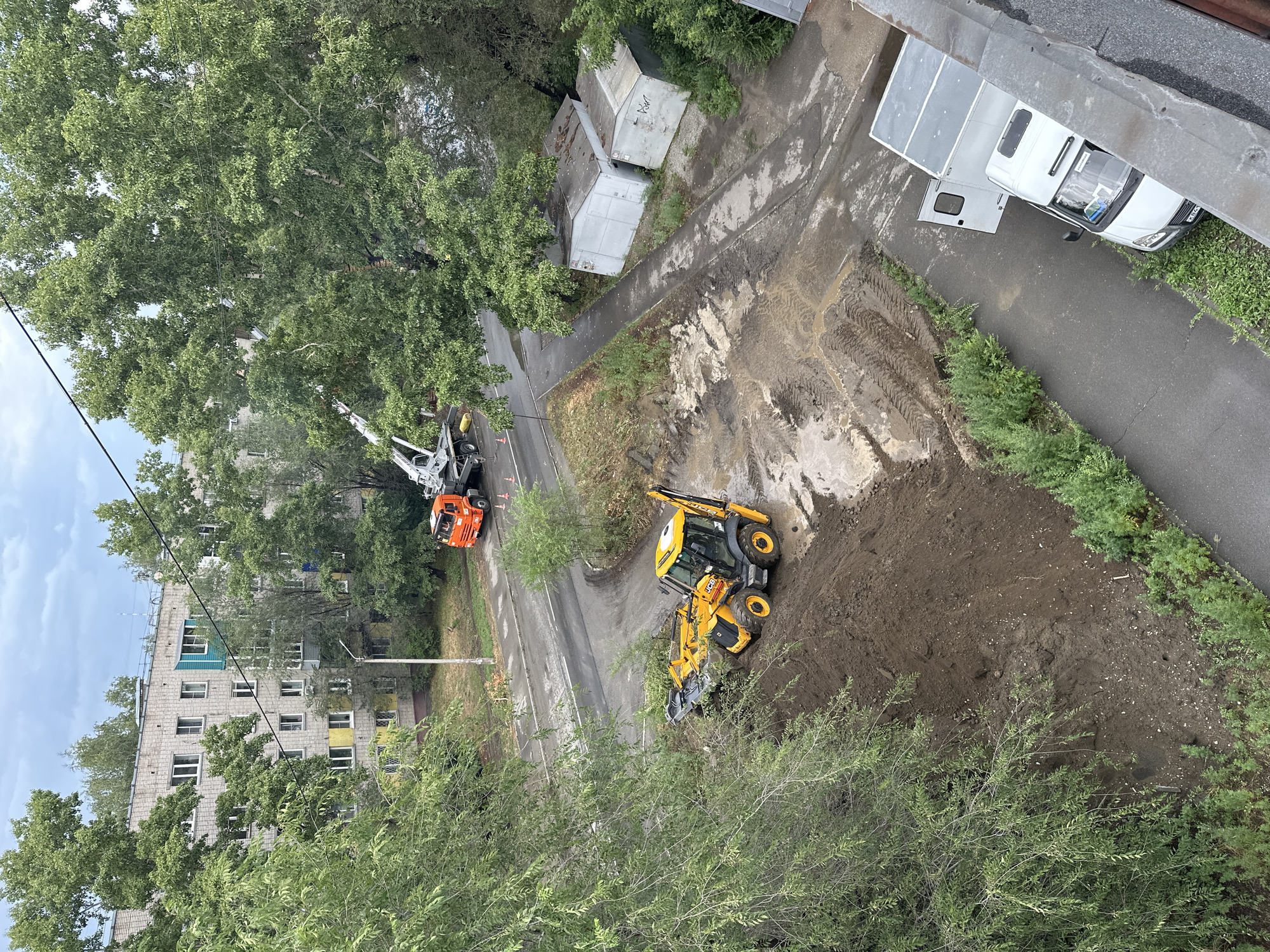 In a residential area of Biysk, Russia, a construction site is visible. A yellow JCB backhoe is actively excavating soil, creating a large mound. An orange crane truck is parked nearby, and a white cargo van is also present. Several small, grey sheds with weathered roofs are situated on the right side of the scene. The surrounding environment is characterized by a paved road, dirt tracks, green trees, and vegetation, with a multi-story apartment building in the background. The sky is overcast.