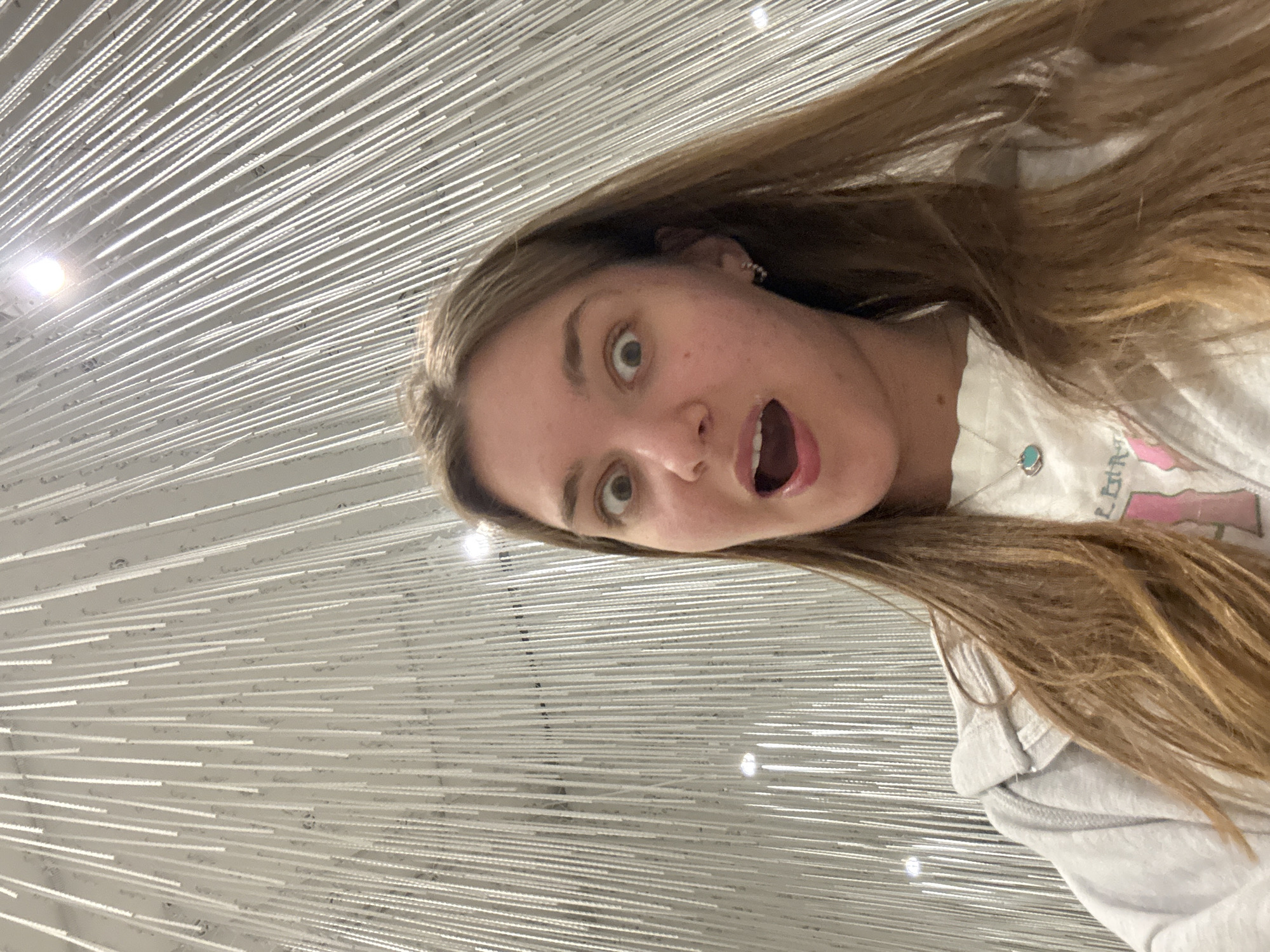 A selfie depicts a fair-skinned woman with long blonde hair. She displays an exaggerated expression of surprise with wide eyes and an open mouth. She wears a casual outfit consisting of a white t-shirt with a pink design and a light gray hooded sweatshirt. The background shows an art installation consisting of numerous thin, white, horizontal rods. The scene is in Perm, Russia.