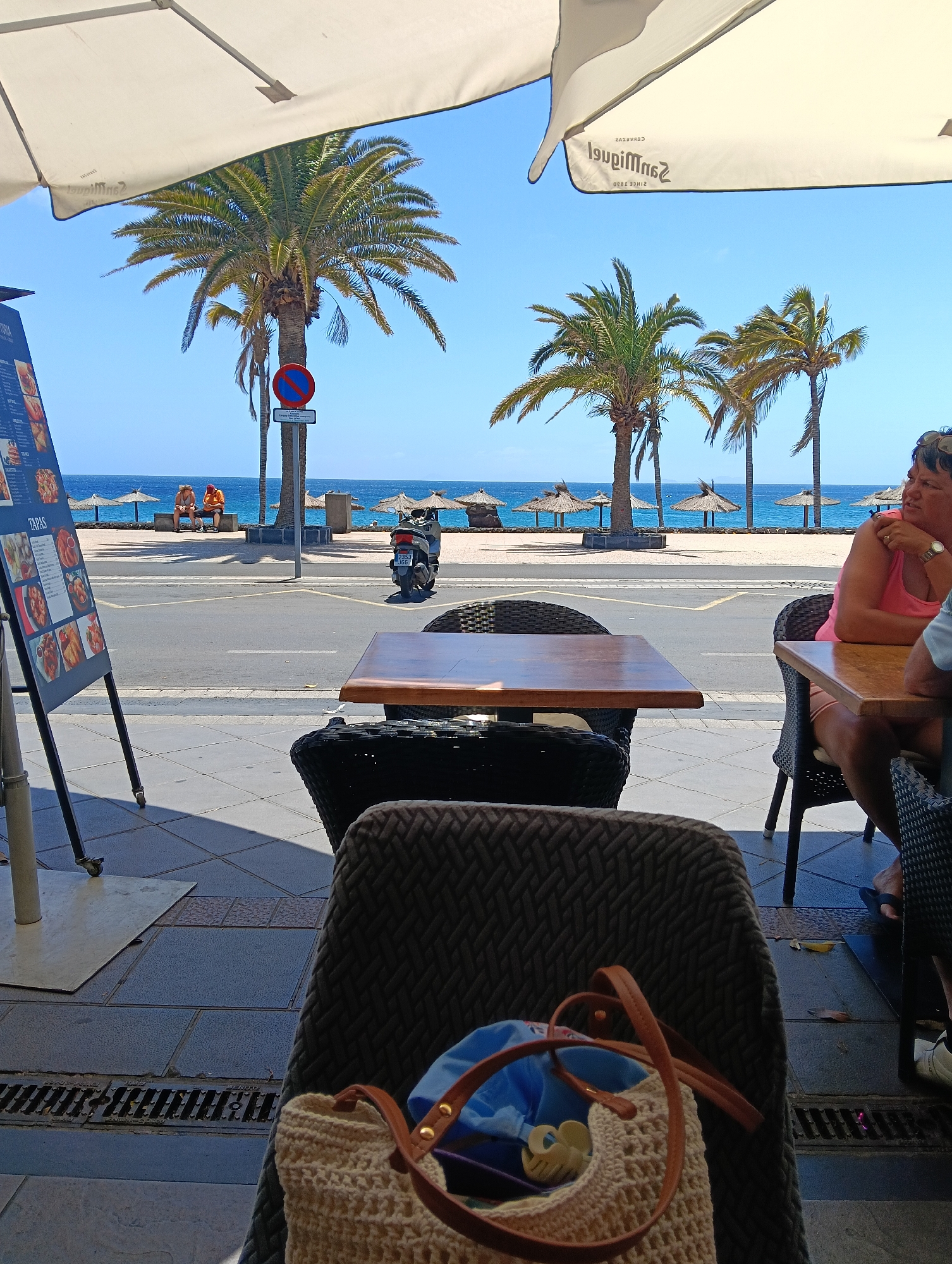 This daytime image captures a vibrant scene from an outdoor cafe in Puerto del Carmen, Spain, looking towards the ocean. In the immediate foreground, a woven chair holds a straw bag containing a blue item and a yellow clip. To the right, a woman in a pink top is seated at a table, looking out towards the sea. Another table is visible directly in front.
To the left, a menu board displays "TAPAS" and various food items. Above, a large white umbrella, partially visible, has the reversed text "San Miguel CERVEZAS SINCE 1890" printed on its underside.
Across the paved street, several tall palm trees stand, with a red and blue "no parking" sign attached to one. A scooter with license plate "2353 JGY" is parked on the road. Beyond the street, a sandy beach is dotted with several thatched umbrellas. Two people are seated on a bench closer to the beach, observing the view. The deep blue ocean extends to the horizon under a clear, bright blue sky, indicating sunny weather. The scene conveys a relaxed, leisurely atmosphere. Shadows on the ground suggest it is mid-morning or afternoon. A drain on the ground shows partially legible text "DEN...".