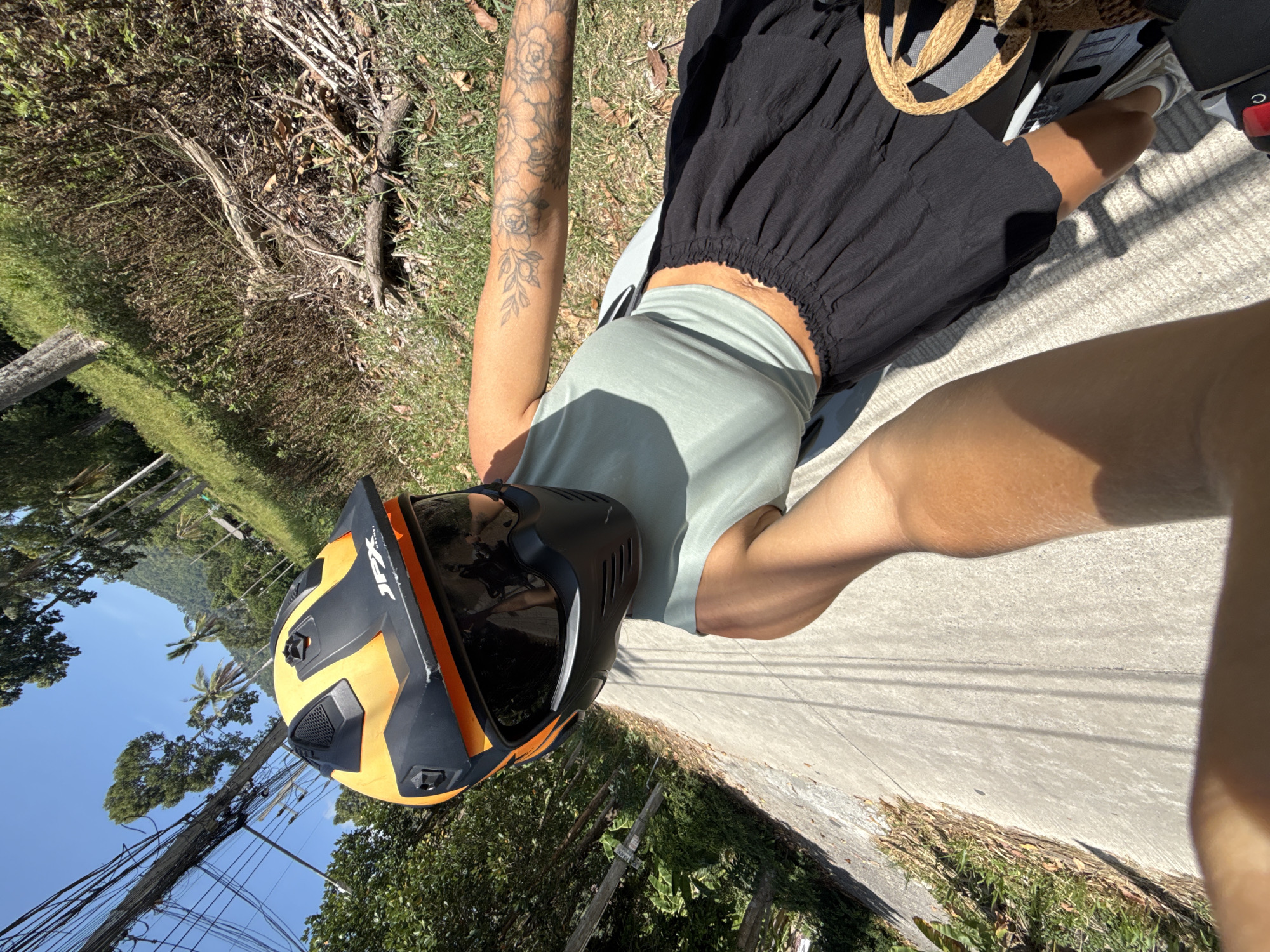 A woman wearing a turquoise tank top, black skirt, and an orange and black helmet is riding a motorcycle on a paved road in Ko Samui District, Thailand. The road is lined with lush green vegetation and trees. Overhead, power lines stretch across the frame. The woman has tattoos on her left arm. The sun is shining, casting shadows on the road. A woven bag is visible on the back of the motorcycle.