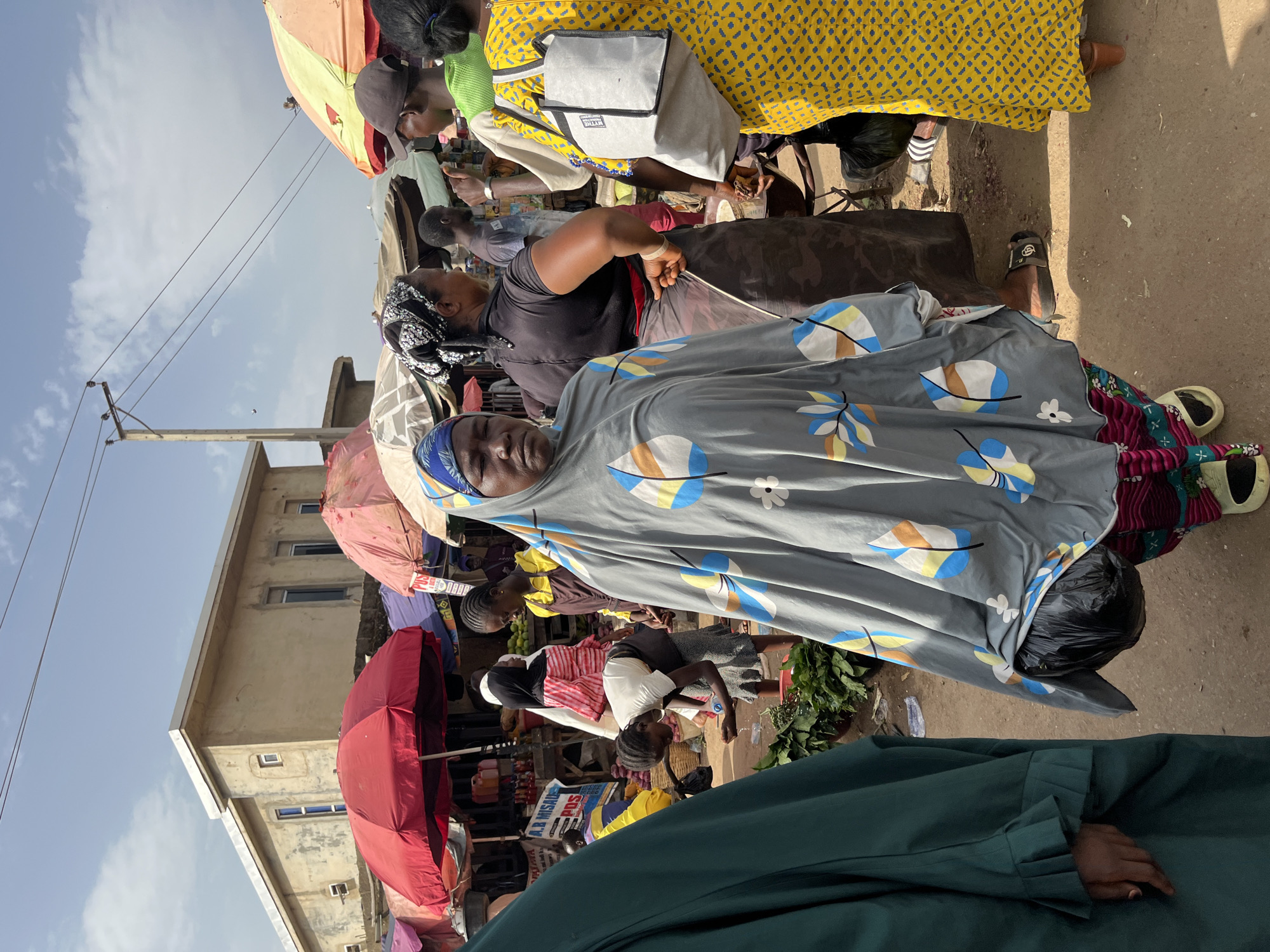 The image depicts a bustling market scene, likely in Rafin Par, Nigeria, during the daytime. Several individuals are present, including women adorned in colorful traditional clothing and headscarves, and children. The setting is outdoors, with stalls and umbrellas providing shade from the sun. Items such as produce, possibly vegetables and fruits, are visible, suggesting the market is a place for commerce. One woman in the foreground wears a gray headscarf with a leaf pattern. Behind her, another woman in a black top is partially obscured. Further back, a woman in a bright yellow patterned dress is visible. Umbrellas of red and pink hues are set up, offering protection from the elements. A building with windows can be seen in the background. The overall atmosphere appears lively and active, characteristic of a busy marketplace. The image is taken in daylight, as indicated by the bright illumination and shadows.