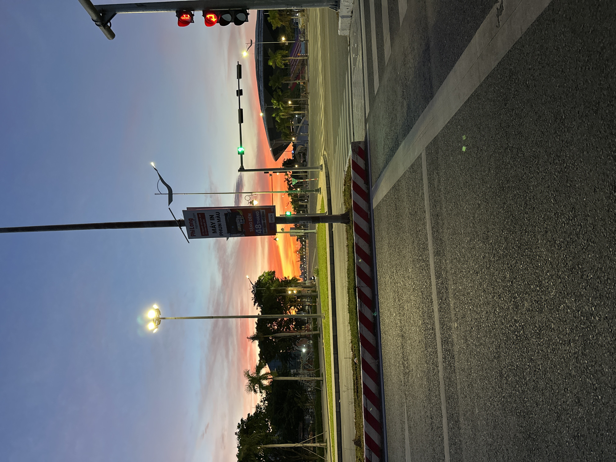 This is a photograph taken at a street intersection in Da Nang, Vietnam, during sunset. The sky displays vibrant hues of orange, pink, and purple, indicating the sun is low on the horizon. The foreground shows a paved road with white lane markings and a pedestrian crossing. To the left, there is a concrete median with red and white striped barriers. A tall lamppost with several bright lights stands prominently in the middle ground. Adjacent to it is a utility pole with a sign that appears to be an advertisement, featuring text in Vietnamese and an image of a printer. Across the intersection, a traffic light shows a green signal. Beyond the road, there are palm trees and other vegetation, with a large, modern-looking building with a distinctive roofline in the background. The overall scene captures a blend of urban infrastructure and natural twilight beauty.