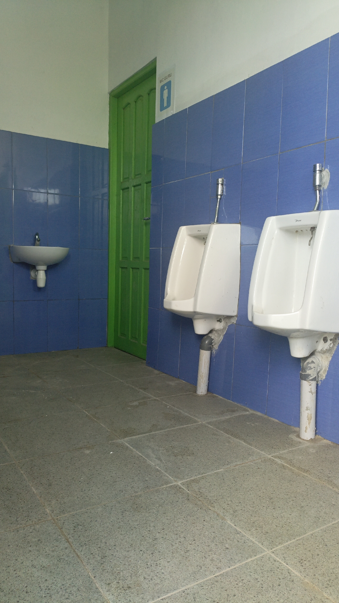This image depicts a men's restroom with blue tiled walls and gray tiled flooring, located in Saring Sungai Binjai, Indonesia. The restroom contains two white ceramic urinals mounted on the blue wall, with visible plumbing pipes extending downwards. To the left, a white ceramic sink is mounted on the wall above a visible plumbing pipe. A bright green door is situated in the corner between the sink and the urinals. A sign with a male silhouette and the text "WC GURU" is mounted on the wall above the door. The environment appears to be clean and functional, with no people visible in the frame.
