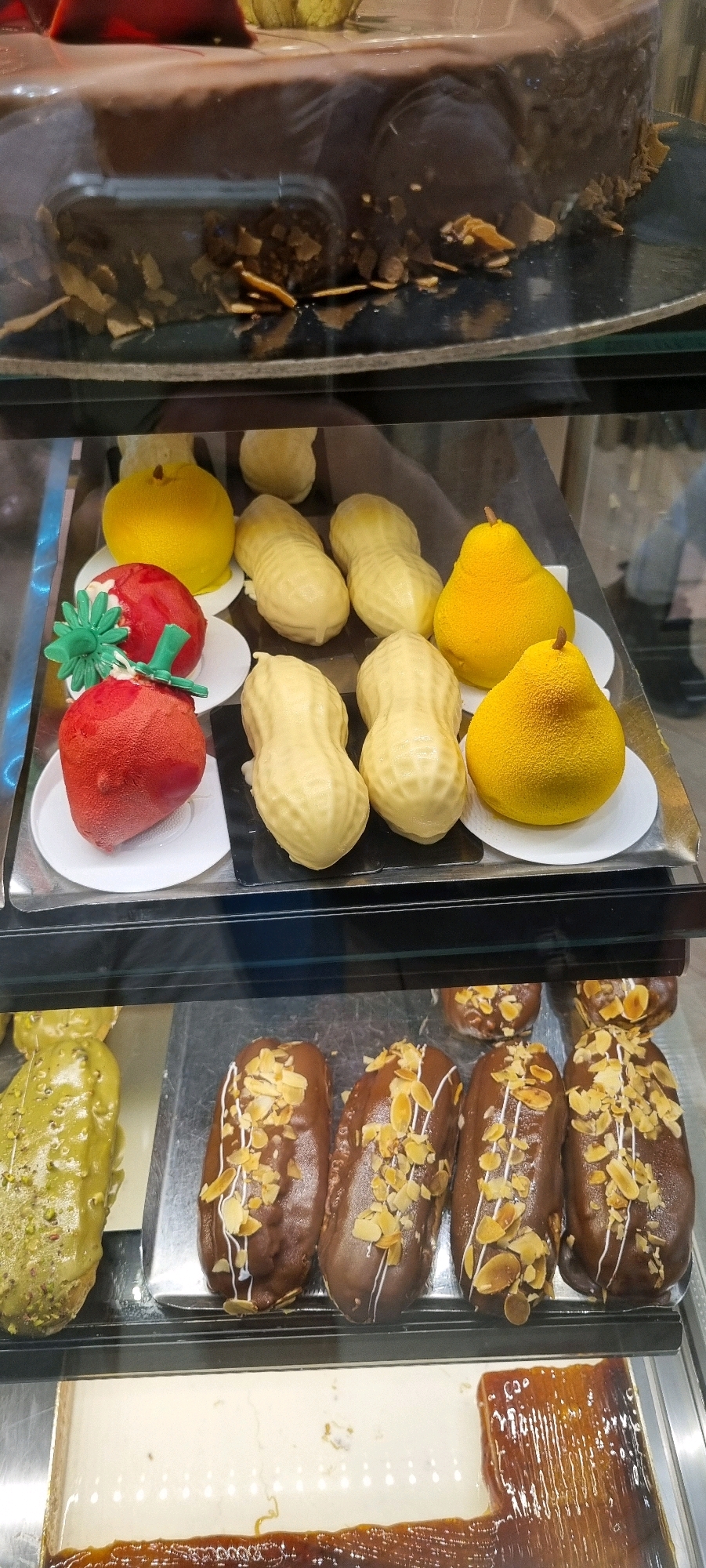 The image shows a display case filled with various desserts. In the top shelf, a large chocolate cake with almond slivers is partially visible. The middle shelf features several individually displayed desserts, including two bright yellow pear-shaped pastries, two red strawberry-shaped pastries with green stems, and multiple light-colored, peanut-shaped pastries. The bottom shelf displays a tray of eclairs covered in chocolate and topped with sliced almonds and white icing, as well as a large rectangular dessert with a glossy caramel topping. The overall environment appears to be a bakery or patisserie. There are no people or specific city locations identifiable in the image.