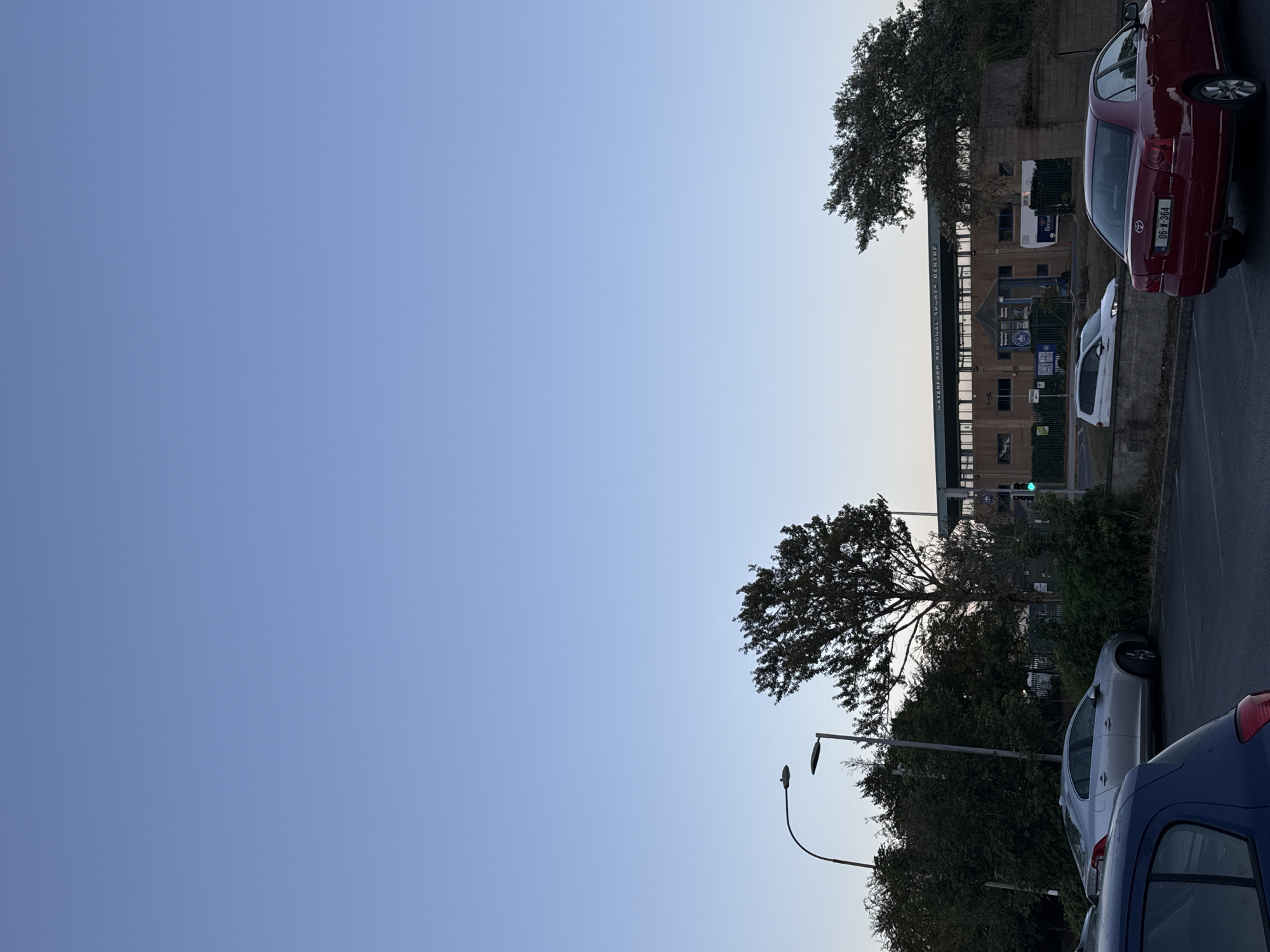 The image captures the exterior of the Waterford Regional Sports Centre in Waterford, Ireland, during the late afternoon or early evening. The sky is a clear, pale blue, indicating a serene weather condition. In the foreground, several cars are parked on an asphalt lot. A silver sedan is parked in the middle ground, angled slightly away from the viewer. To the right, a red sedan is parked, facing the viewer's left. A portion of a blue car is visible in the lower left corner.
The sports centre building itself is a prominent structure with a light-colored facade and dark-colored trim. A sign above the building's entrance reads "WATERFORD REGIONAL SPORTS CENTRE." The building appears to be a modern, multi-story facility with numerous windows.
Surrounding the building and parking lot are various trees and shrubbery, contributing to a naturalistic setting. Two streetlights are visible on the left side of the image, their dark poles and lamps contrasting with the sky. A green traffic light is illuminated on a pole near the sports centre entrance, suggesting an active roadway or pedestrian crossing nearby. The overall scene conveys a sense of a public facility in a suburban or semi-urban environment.