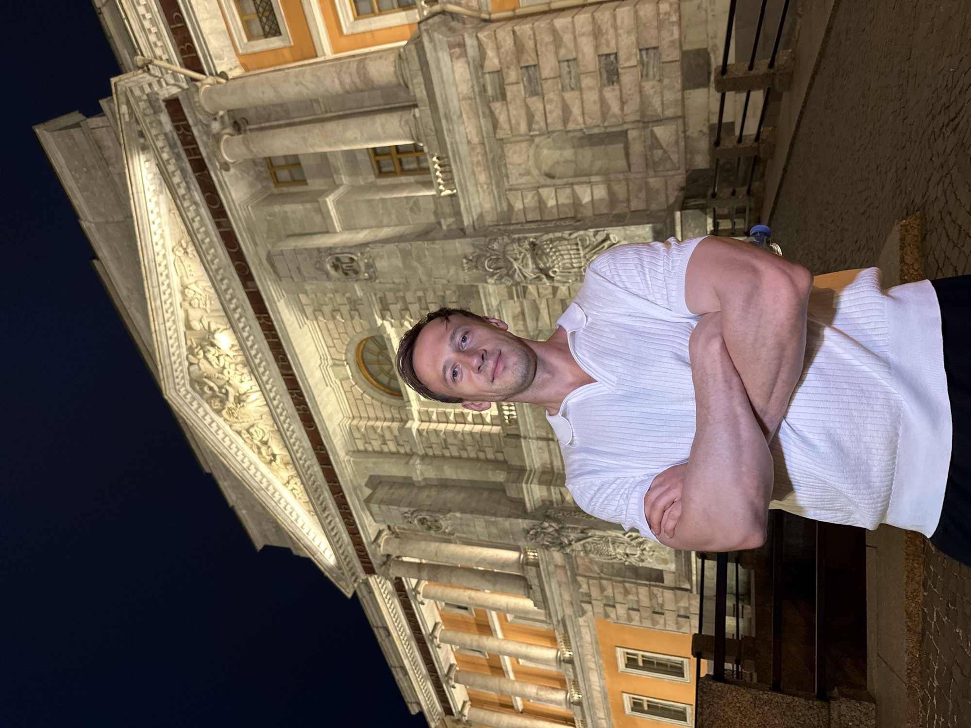 A man stands with his arms crossed in front of a large, ornate building at night. The building features classical architecture with columns, intricate carvings, and illuminated windows. The man is wearing a white polo shirt and dark pants. The ground is paved with cobblestones. The scene appears to be in Saint Petersburg, Russia, likely in front of the General Staff Building on Palace Square, due to the building's distinctive facade and the historical context of such architecture in the city.