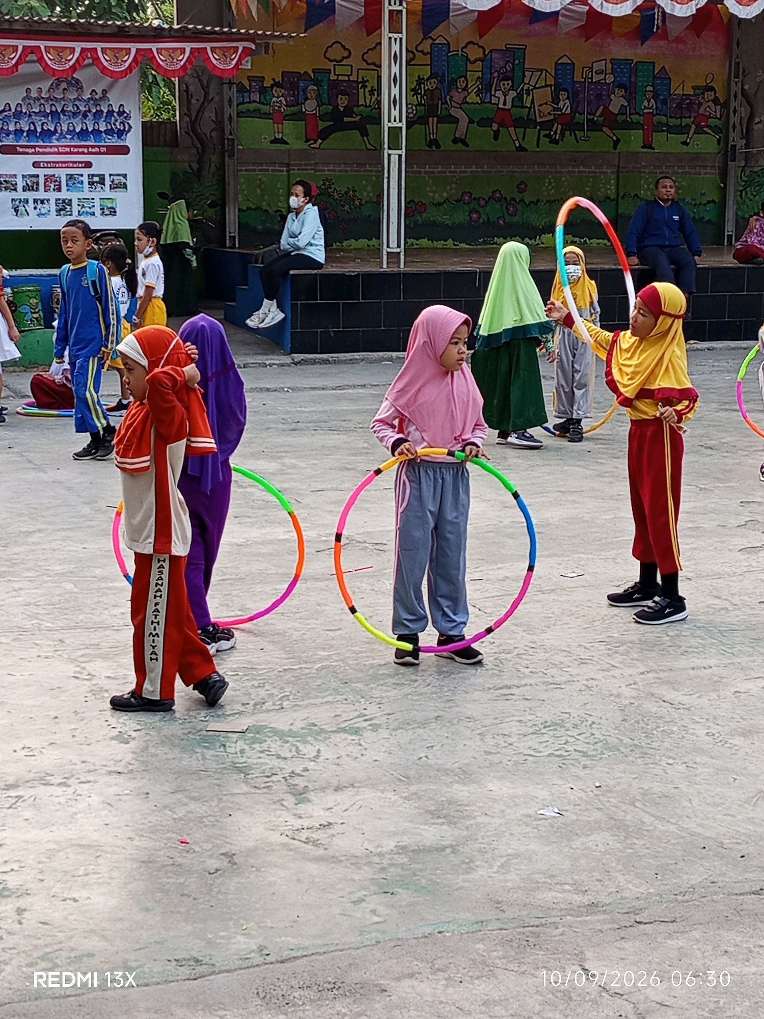 The image depicts an outdoor activity taking place on a paved area, likely a schoolyard or public space in Cikarang, Indonesia. Several children, dressed in school uniforms or athletic wear, are engaged in activities involving hula hoops. One girl in a pink hijab and grey pants holds a hula hoop, while another girl in a yellow hijab and red pants appears to be demonstrating or participating with a hula hoop. A boy in a red and white tracksuit is also visible, as is another child in a purple garment. In the background, there are murals on a wall and a structure that resembles a stage or elevated platform, with adults seated and standing. A banner with text and images is also visible on the left side of the frame. The ground is concrete with some patches of discoloration and debris. The lighting suggests it is daytime, possibly morning or afternoon. A timestamp "10/09/2026 06:30" and "REDMI 13X" are present in the lower right and left corners respectively.