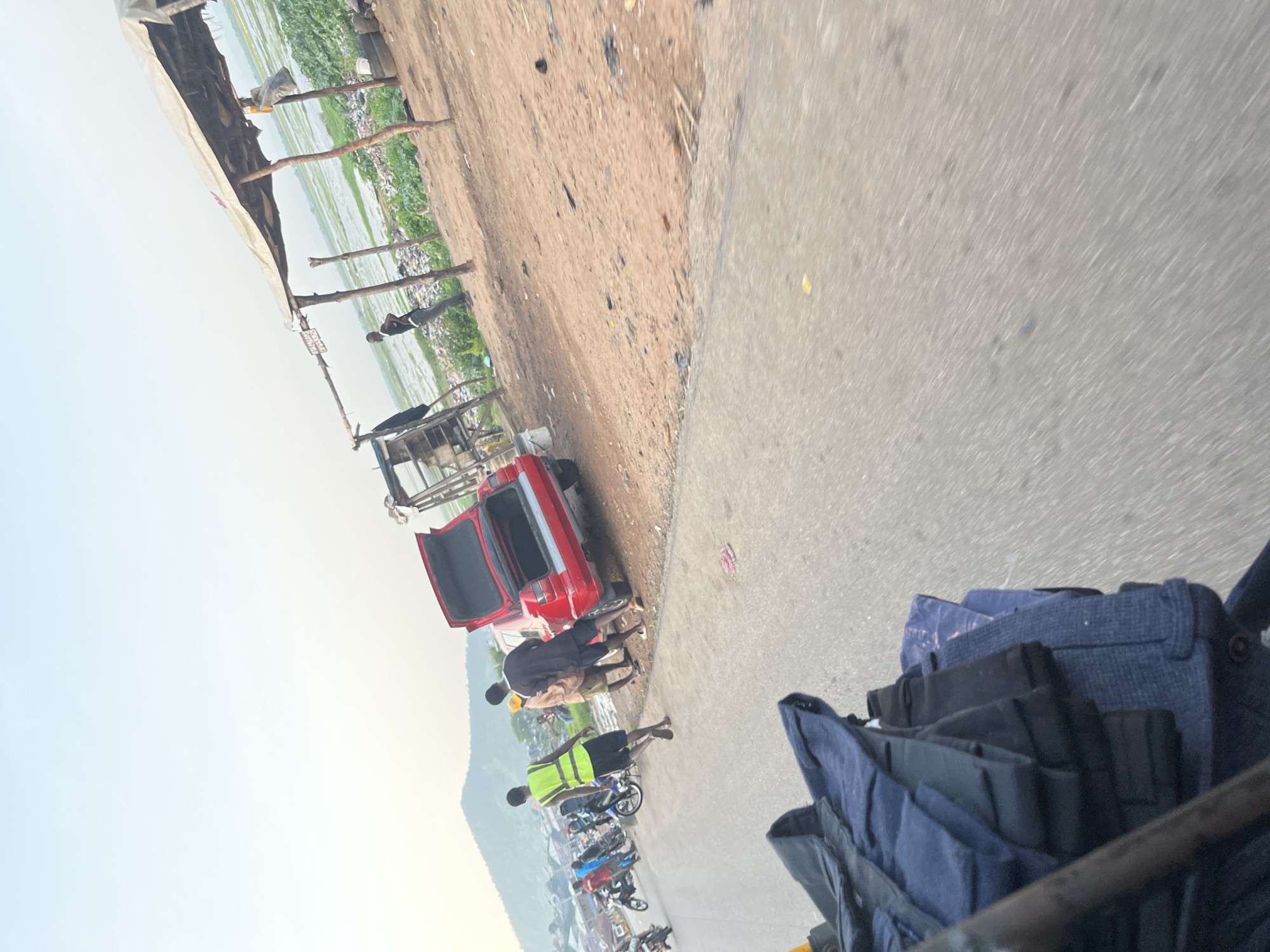 An outdoor scene depicts a road and its surroundings with several individuals and objects. In the foreground, a stack of dark blue bags and garments is visible, suggesting luggage or cargo. The road itself appears paved and is partially occupied by a red car with its trunk open, attracting the attention of a few men. One man is wearing a bright yellow safety vest, while others are dressed in casual attire. Several motorcycles are parked or in motion on the left side of the road, indicating a mode of transportation.
Beyond the road, the environment transitions to a dirt area with a makeshift shelter constructed from wood and a tarp, possibly a roadside stall or resting spot. In the background, a body of water, likely a swamp or marsh, stretches out, with a distant hillside or mountain range. The area around the shelter appears to be littered with debris, including tires and various forms of refuse. The sky is a pale, hazy blue, suggesting either early morning or late afternoon lighting. The overall scene suggests a roadside environment in a developing or rural area.