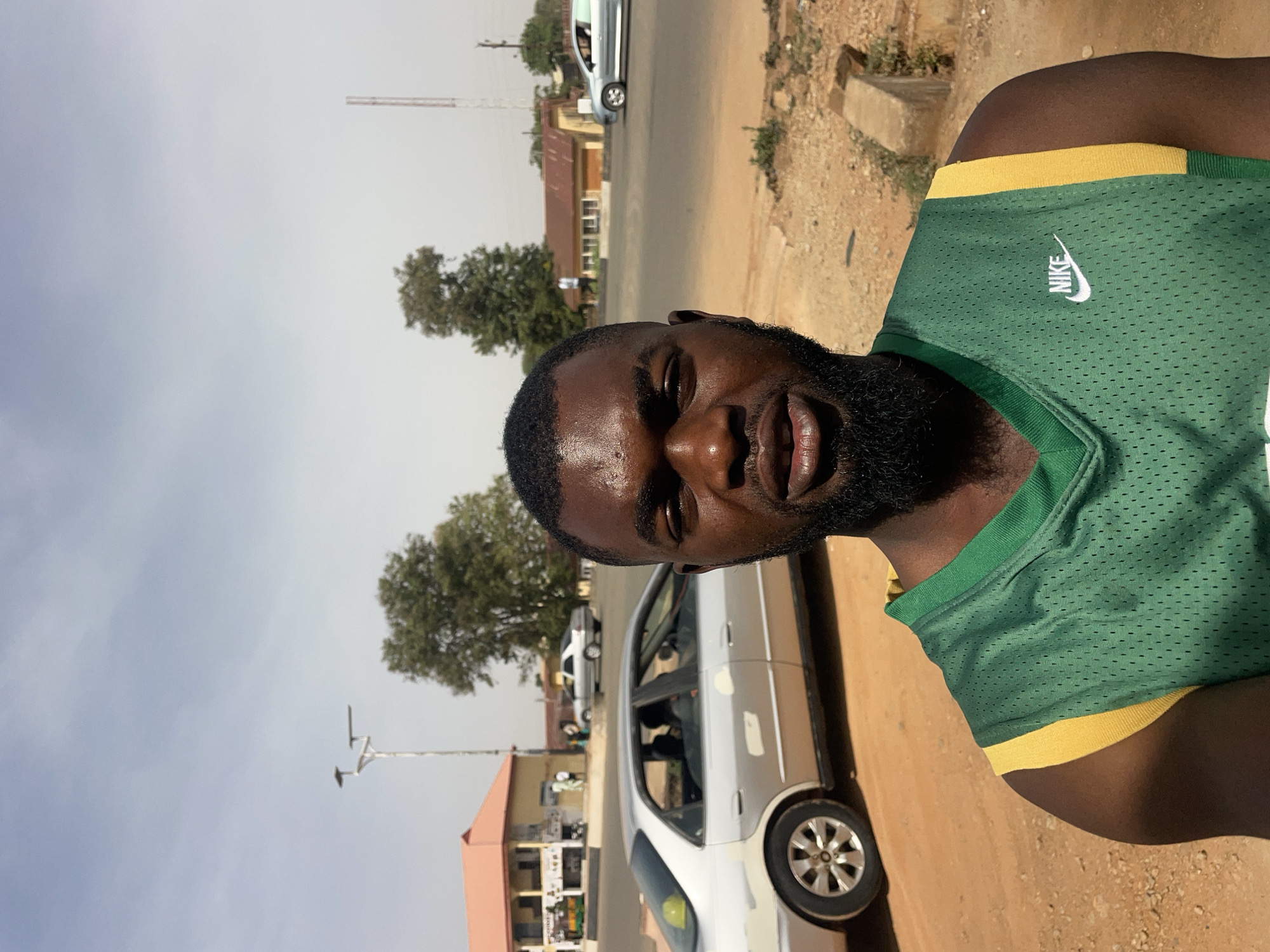 A man with a beard and a green Nike shirt stands in an urban setting. Behind him, there is a street with parked cars and buildings, suggesting a populated area. A tall tower and trees are visible in the background. Based on the information provided, the location is in Jos, Nigeria.