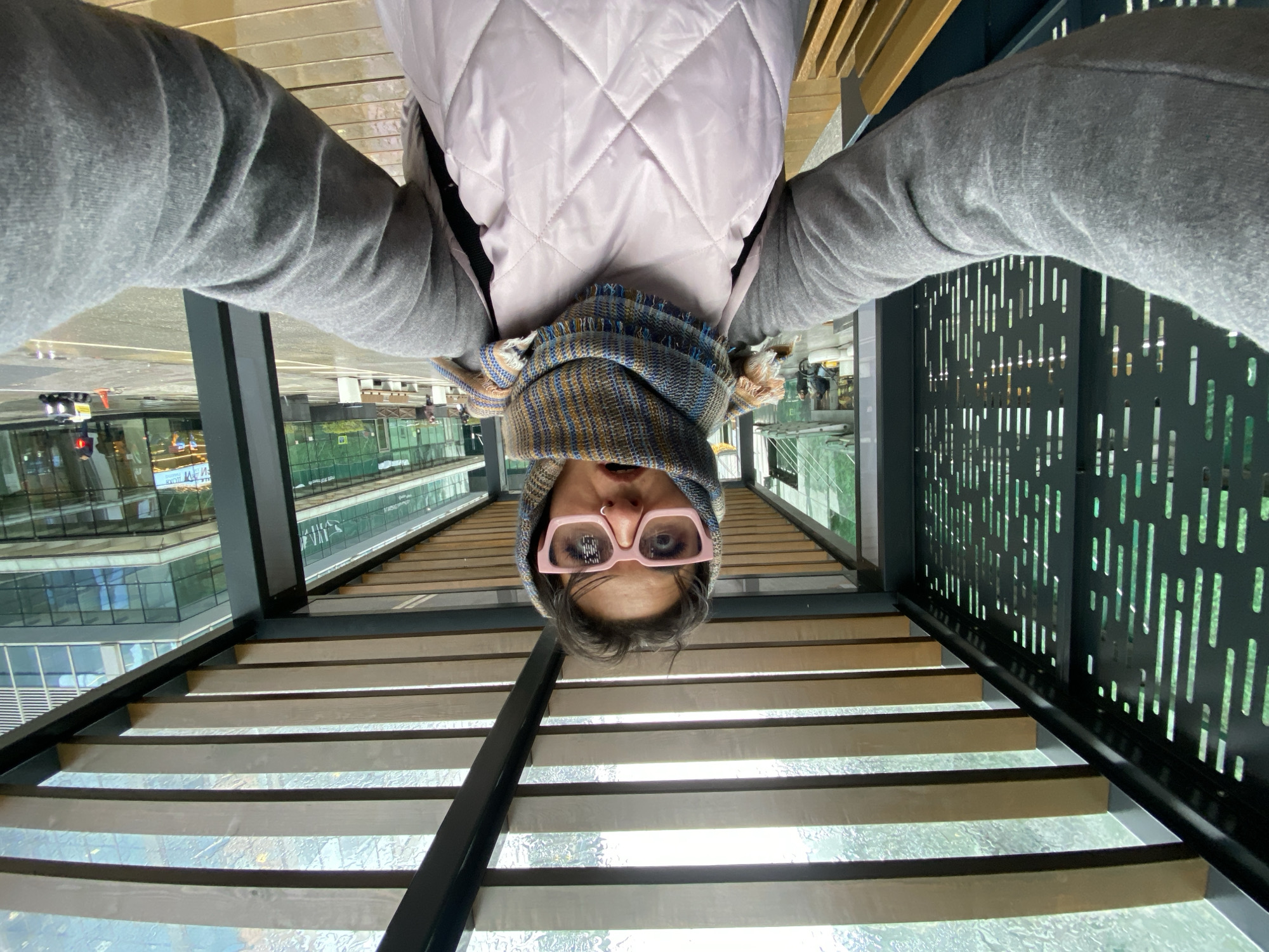 A person wearing pink-framed glasses, a patterned scarf, a grey quilted jacket, and a grey top, is positioned upside down in the frame, suggesting the photo was taken from an overhead or inverted perspective. The background features a glass-bottomed structure, possibly a bridge or walkway, with city buildings visible below. The setting appears to be an urban environment, but further details about its location in Khimki, Russia, are not discernible from the visual data.